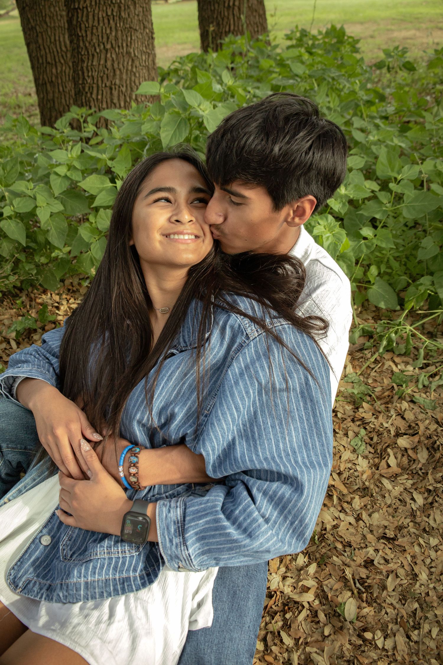A young couple shares a romantic moment in a park, with the man kissing the smiling woman on the cheek.