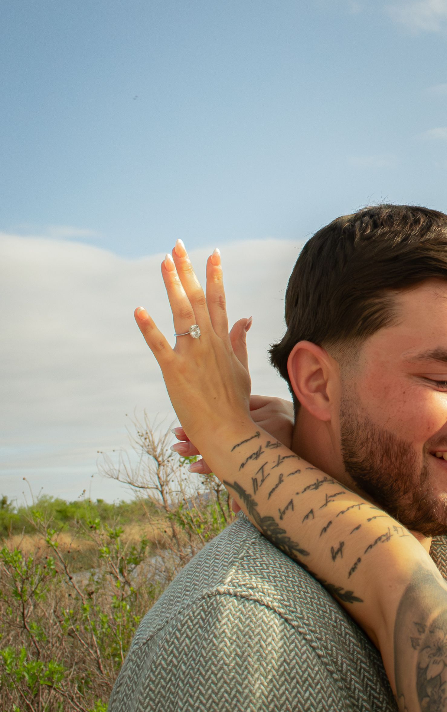 A woman shows off her engagement ring while embracing a tattooed man outdoors under a blue sky.
