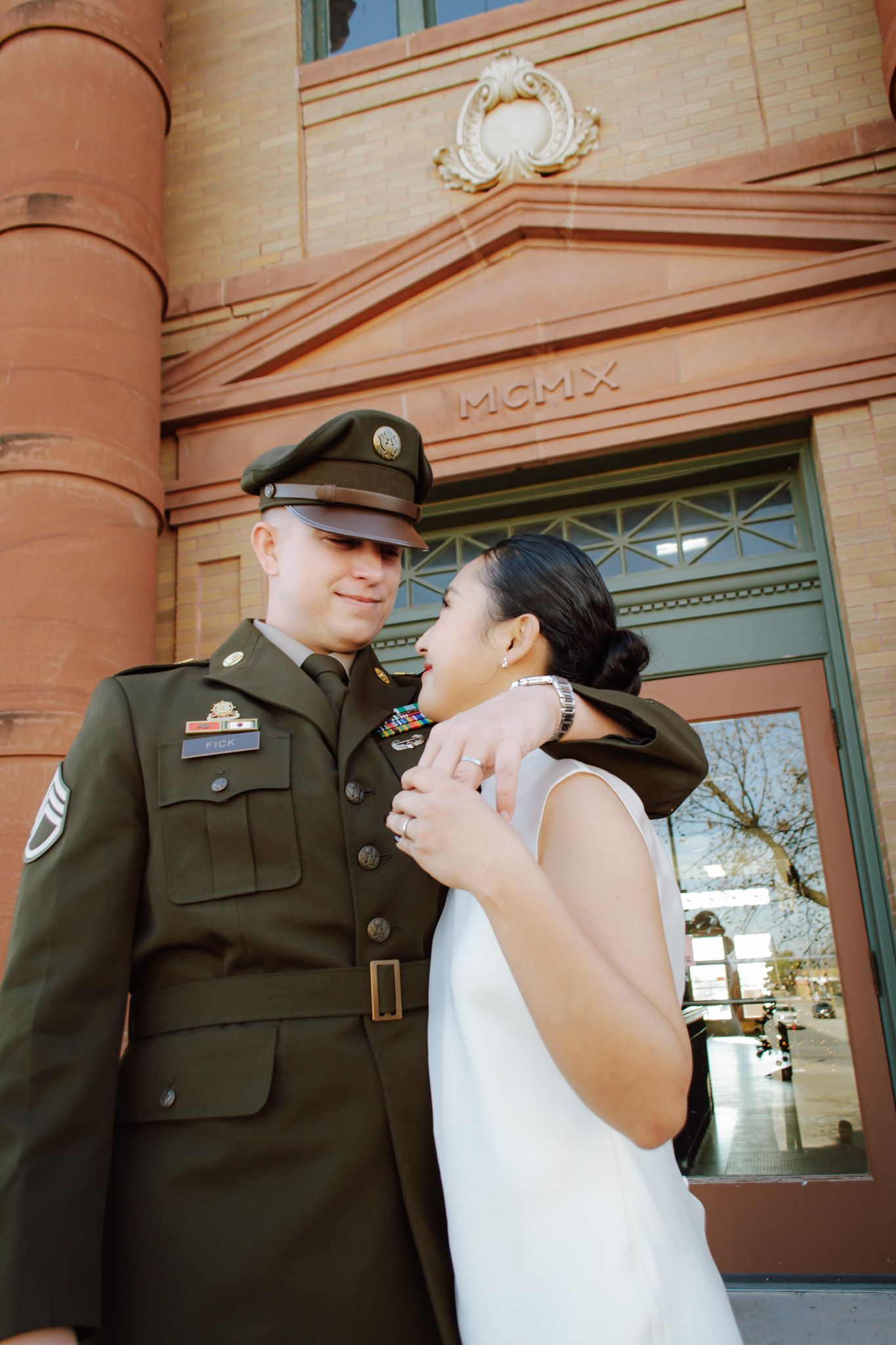 Military groom and bride sharing a tender look outside a historic brick building marked MCMX.