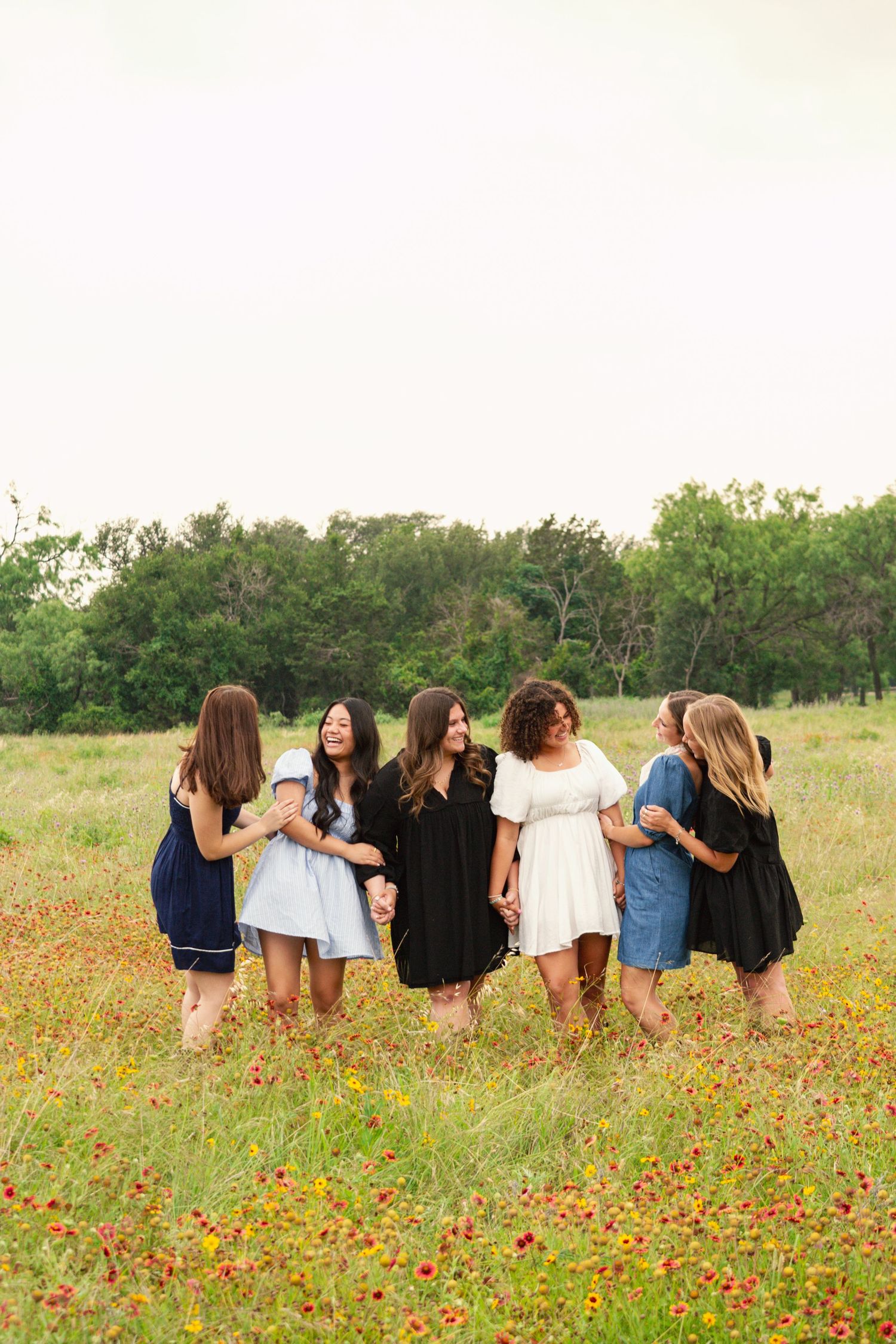 Group of six young women in dresses standing in a circle in a colorful wildflower field.