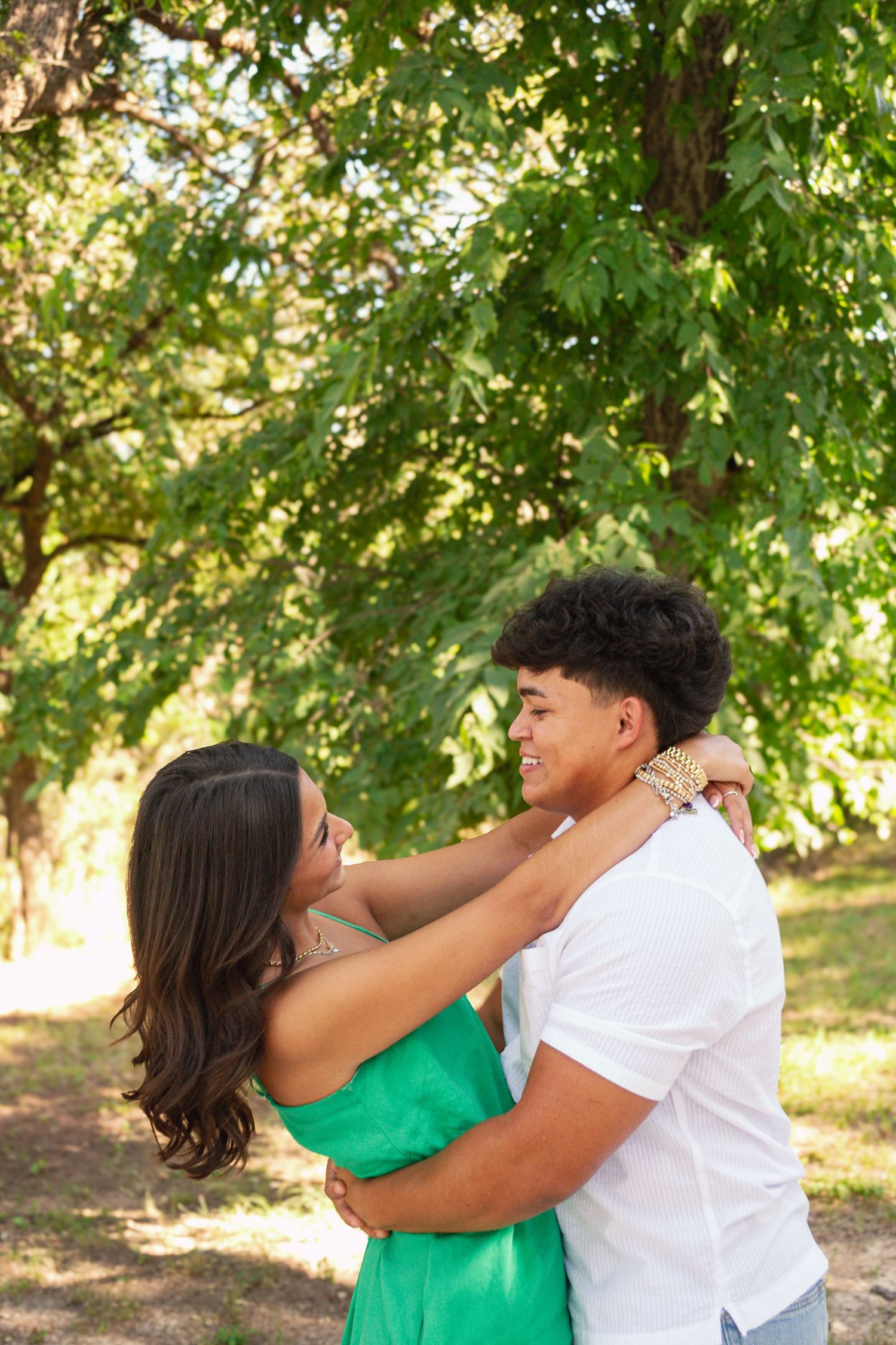 A young couple embraces and smiles at each other outdoors, surrounded by lush green trees on a sunny day.