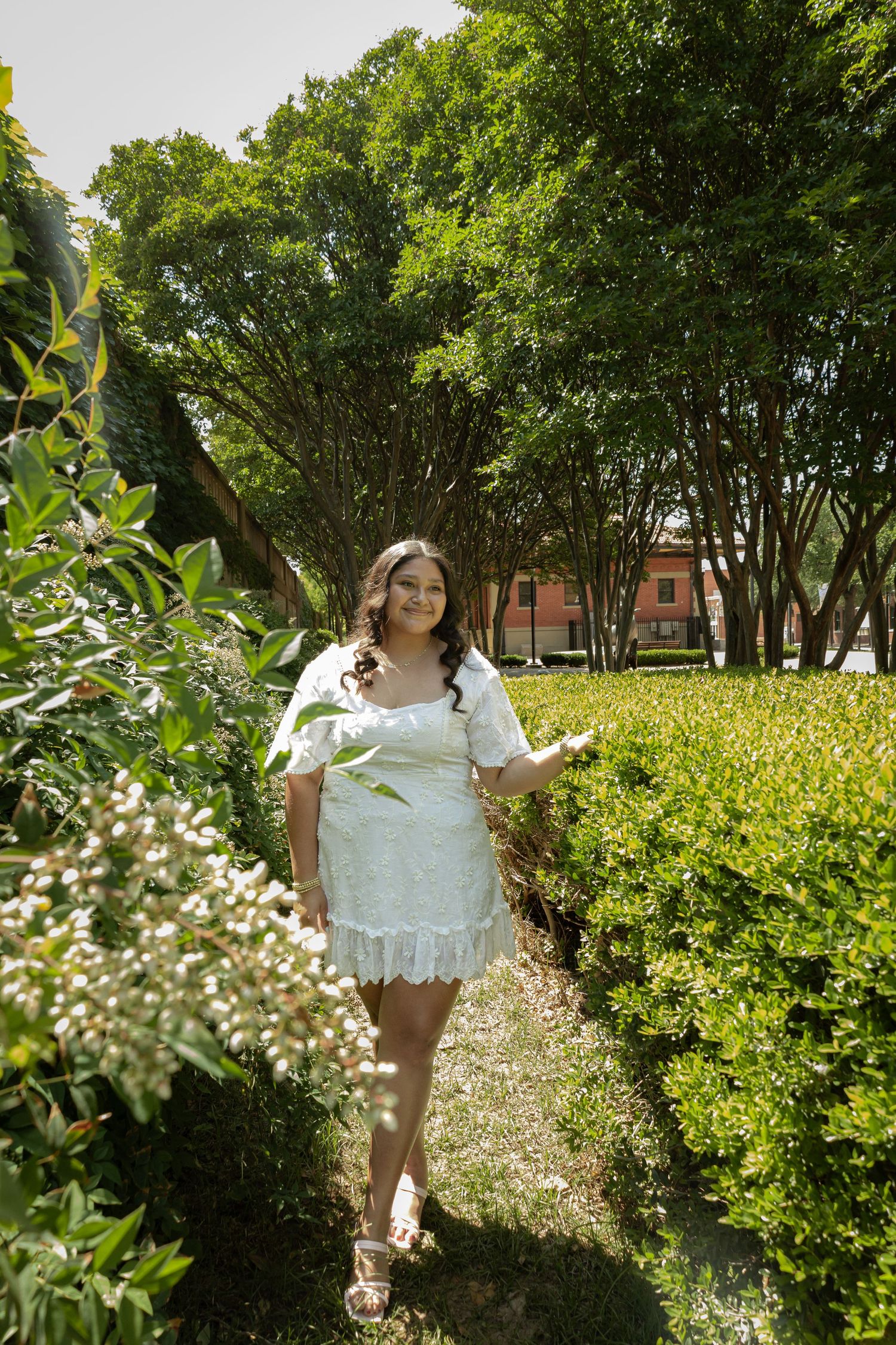 Woman in white lace dress walking through lush green garden path on sunny day.