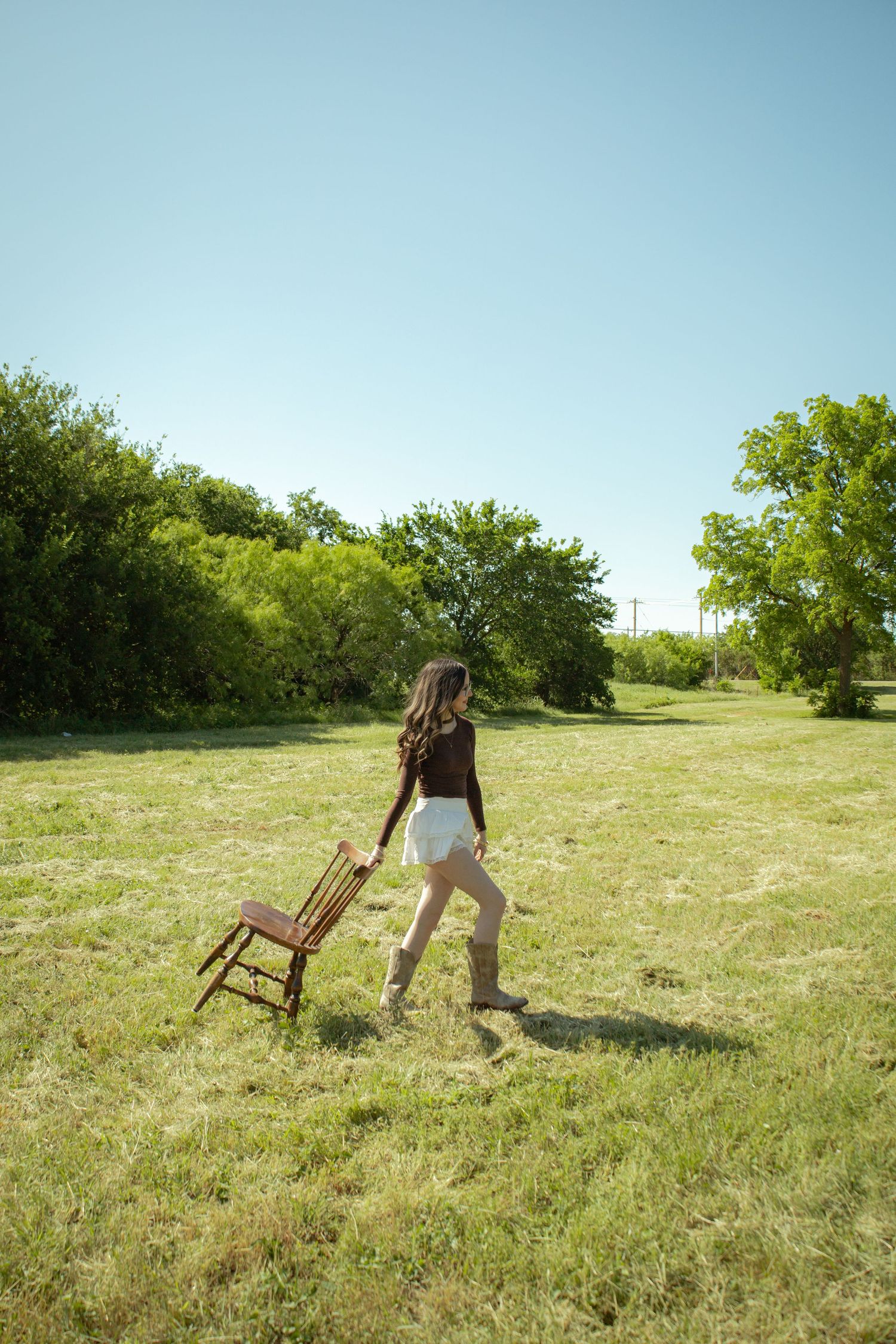 Woman in white shorts and boots carrying a wooden chair across a lush green field on a sunny day.