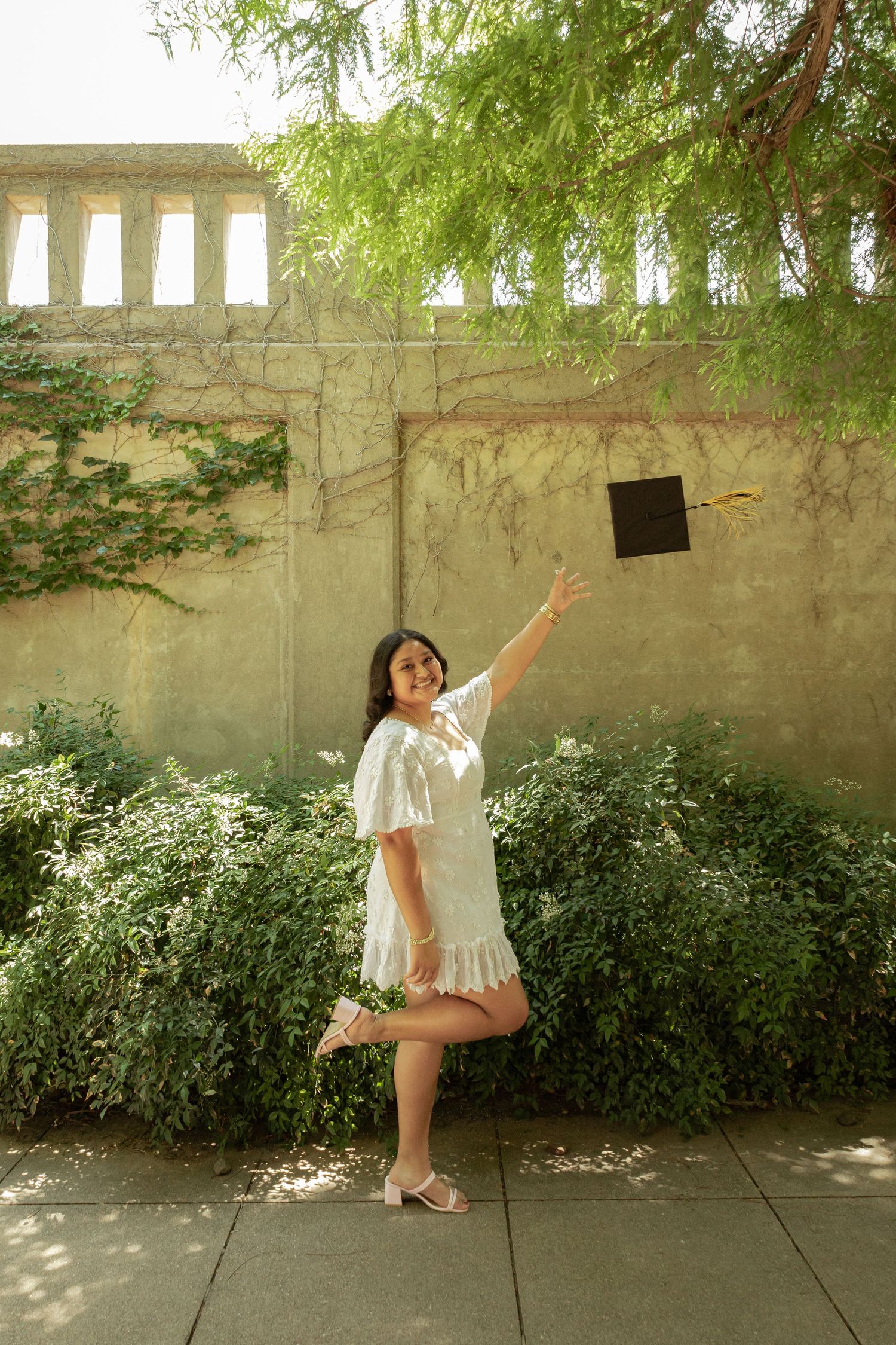 Young woman in white dress tossing graduation cap outdoors near green bushes and stone wall.