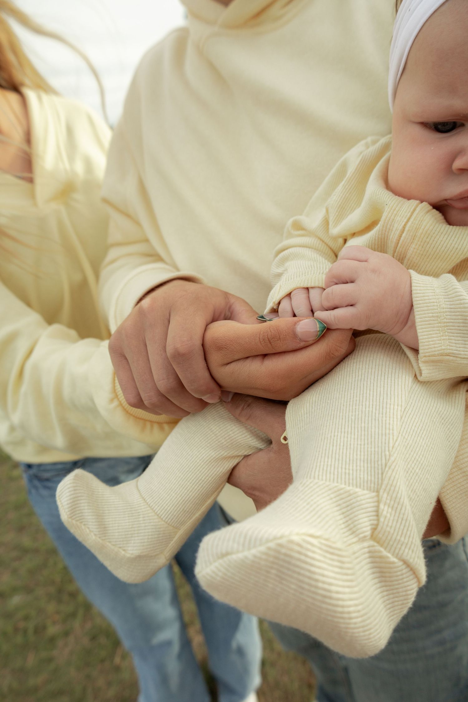 Parents in matching cream hoodies tenderly holding their baby dressed in a yellow ribbed outfit outdoors.