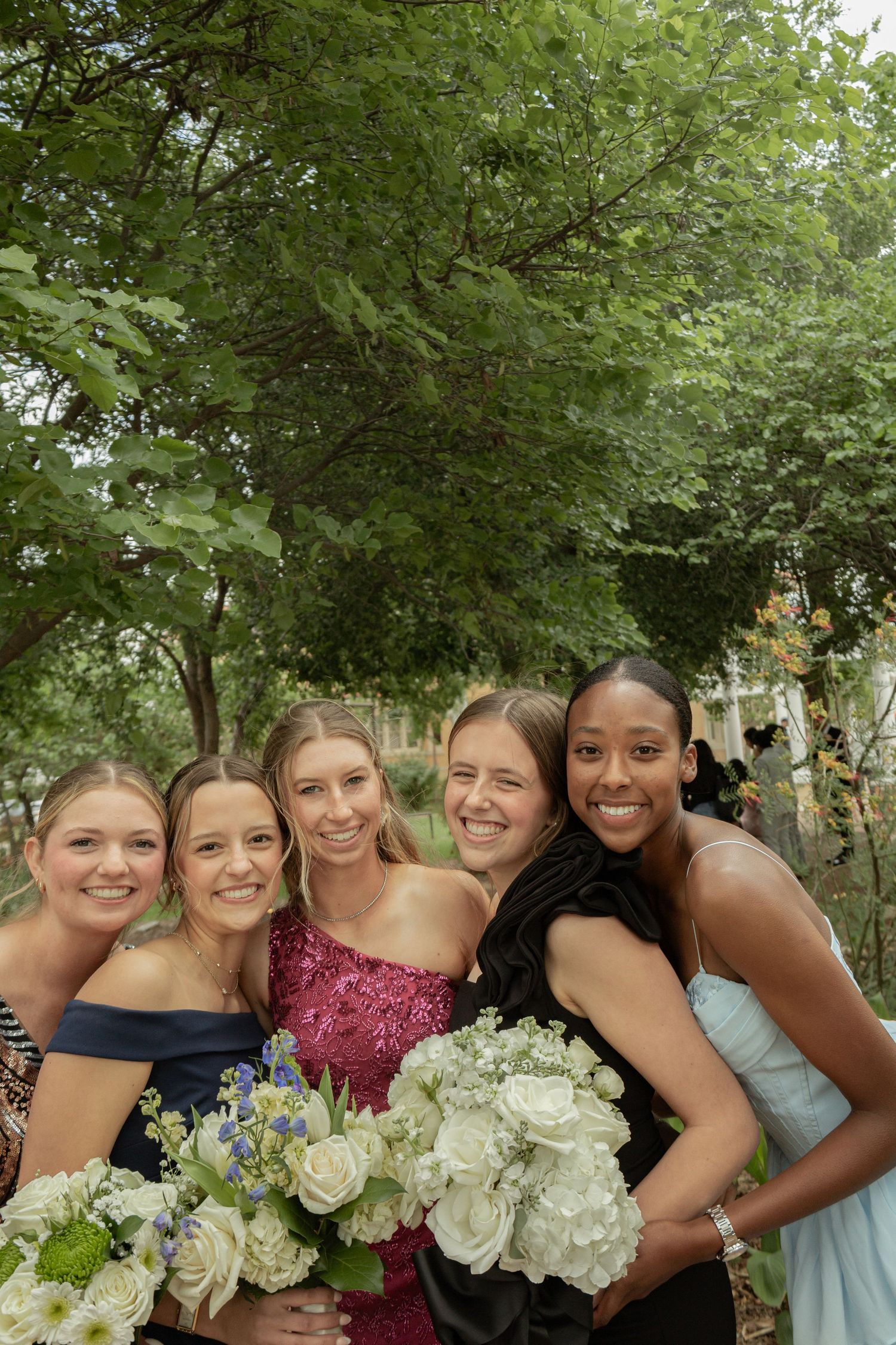 Five young women in formal dresses smile together outdoors holding elegant white flower bouquets.