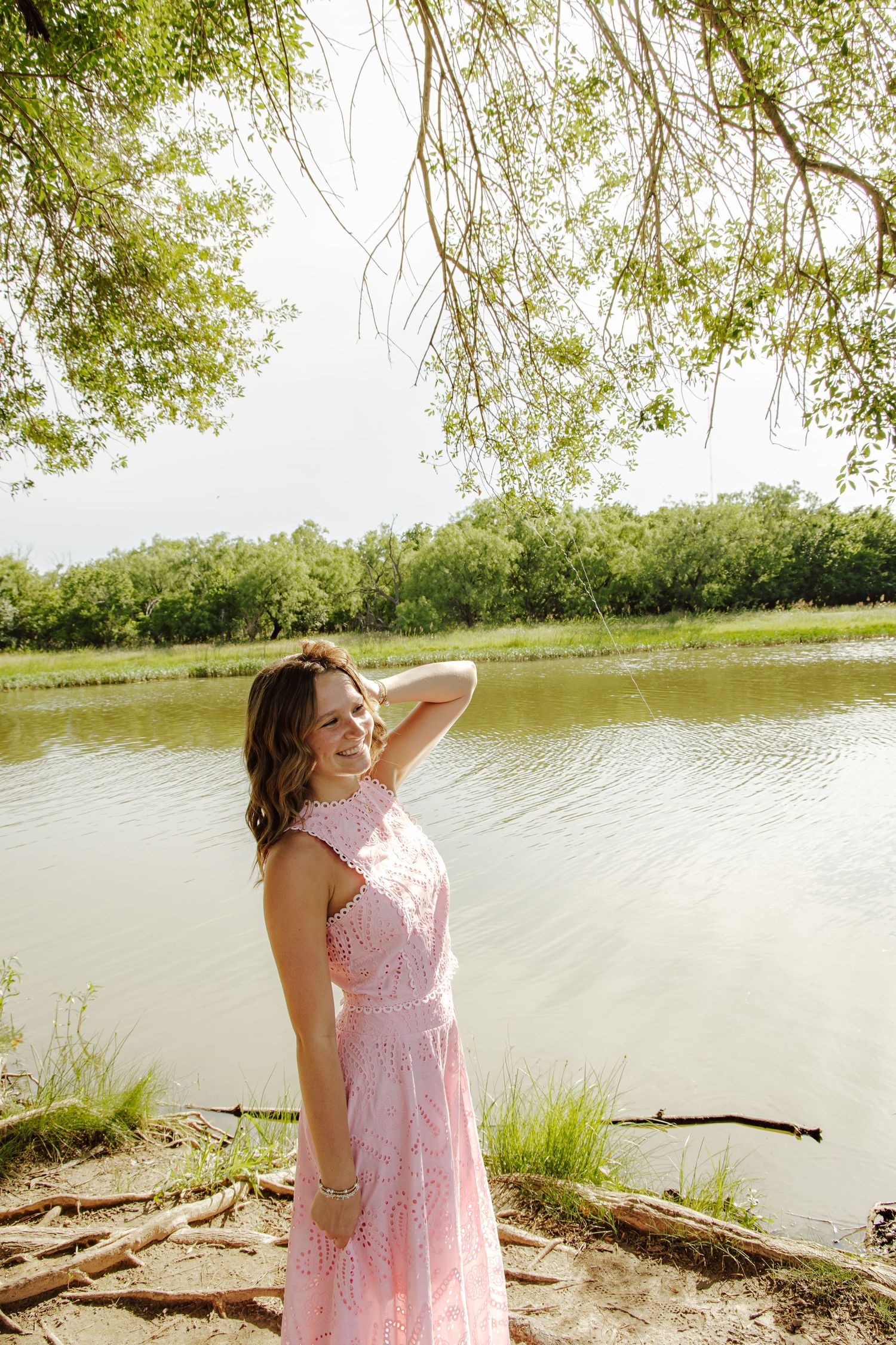 Woman in pink lace dress smiling by a serene river, surrounded by lush green trees on a sunny day.