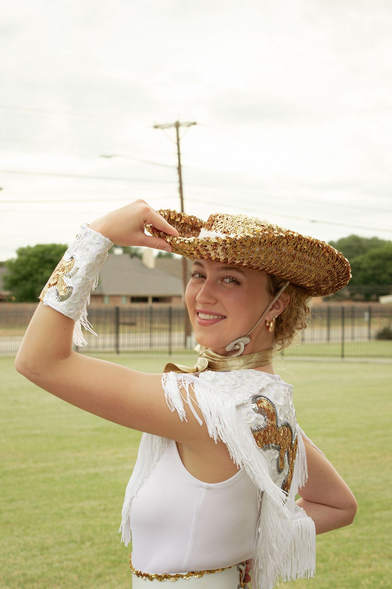 Smiling young woman in white fringe costume and gold sequin cowboy hat posing outdoors on a green field.