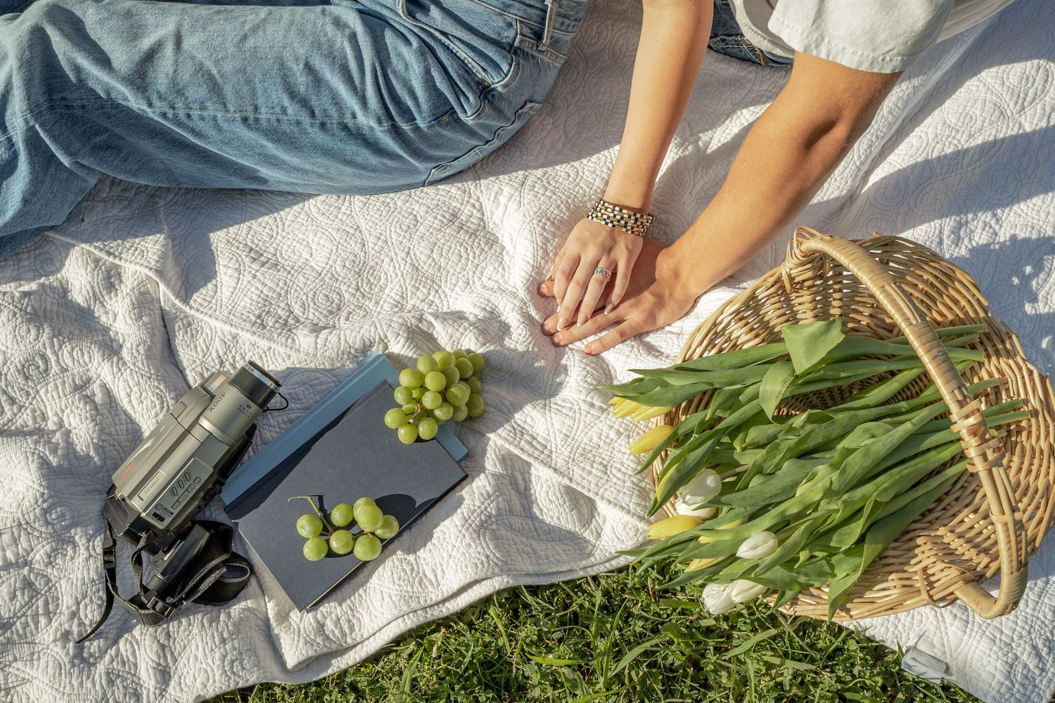 Couple holding hands on a white picnic blanket with grapes, tulips, a camera, and a thermos on a sunny day.