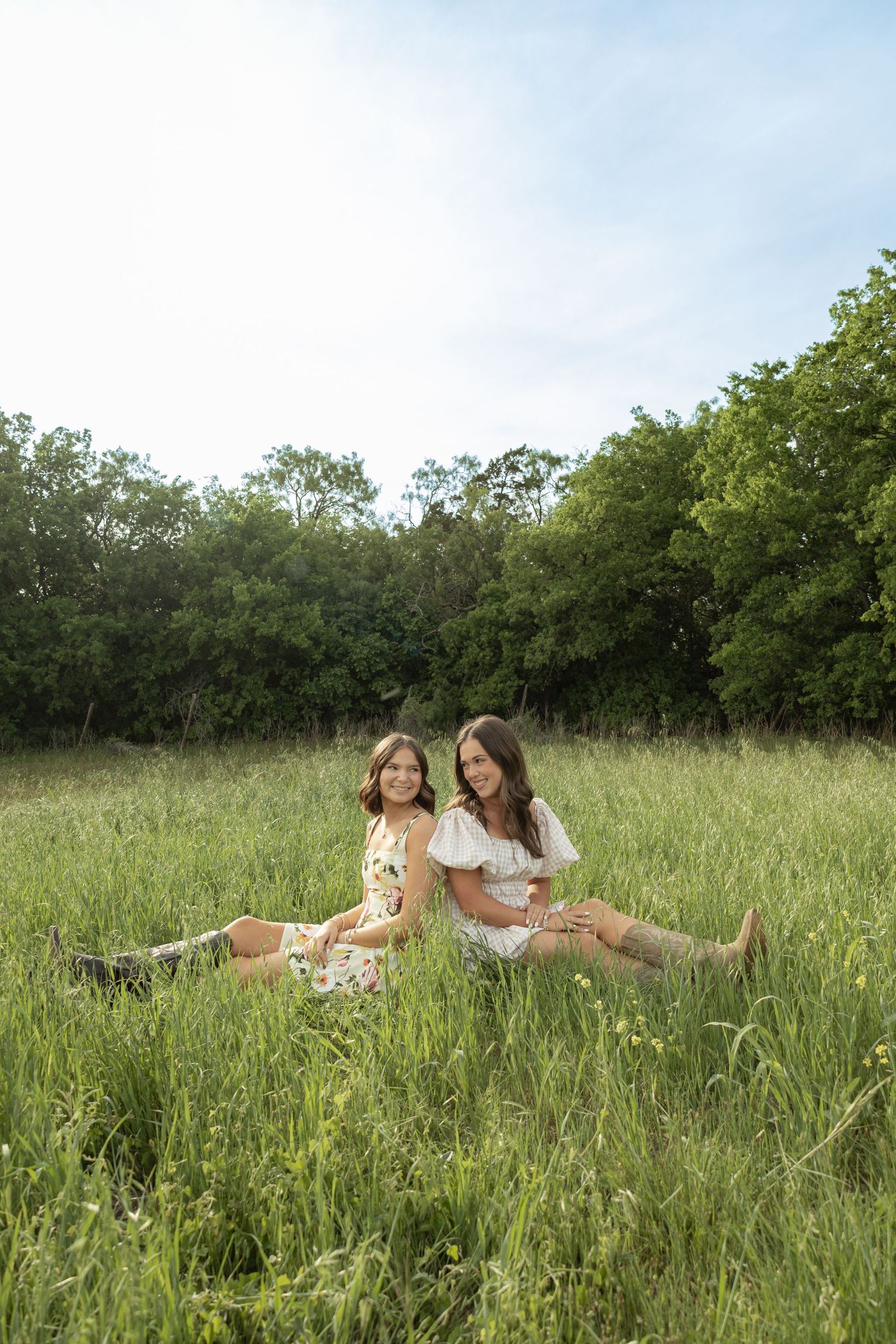 Two women in floral dresses and boots sitting together in a lush green meadow with trees in the background.