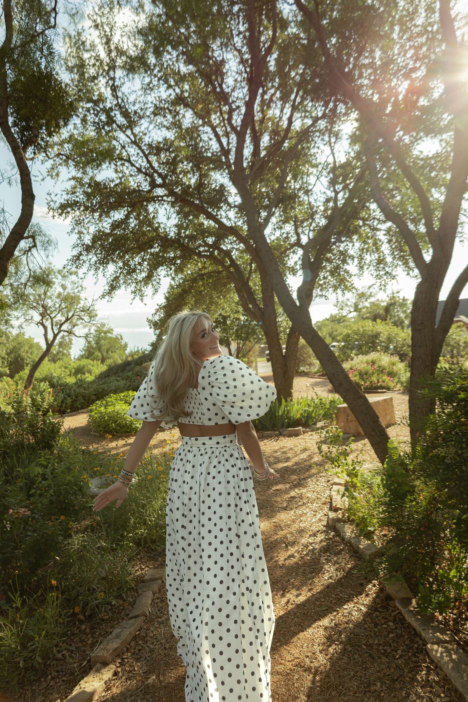 Woman in white polka dot maxi dress walking through sunlit garden path surrounded by lush green trees.