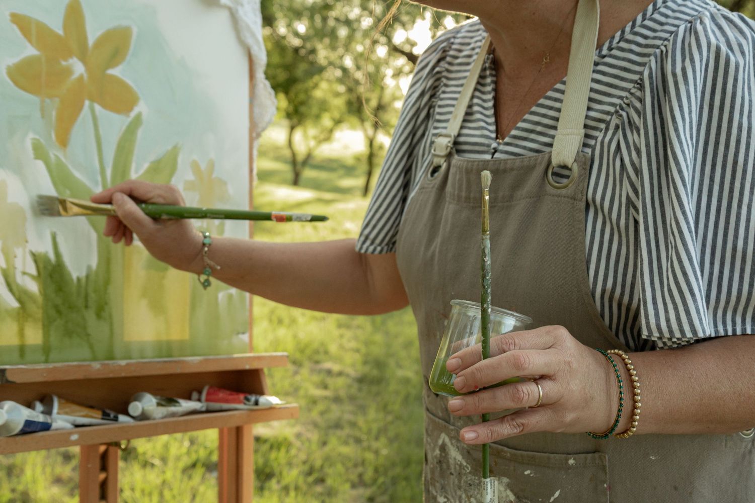 Artist painting floral canvas on outdoor easel, wearing apron and striped shirt, holding paintbrush and glass jar.