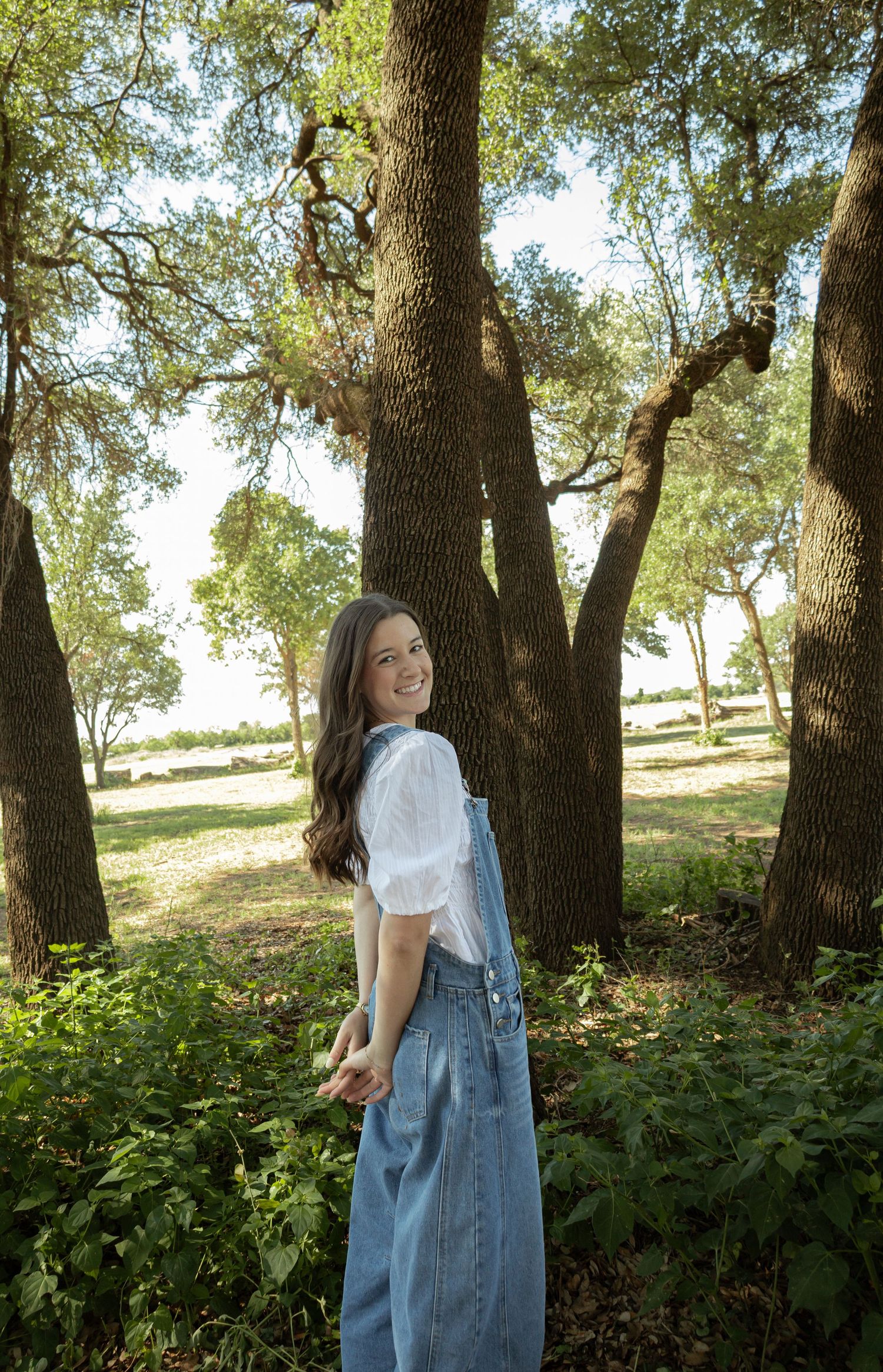 Young woman in white top and denim overalls smiling in a lush green park surrounded by tall oak trees.