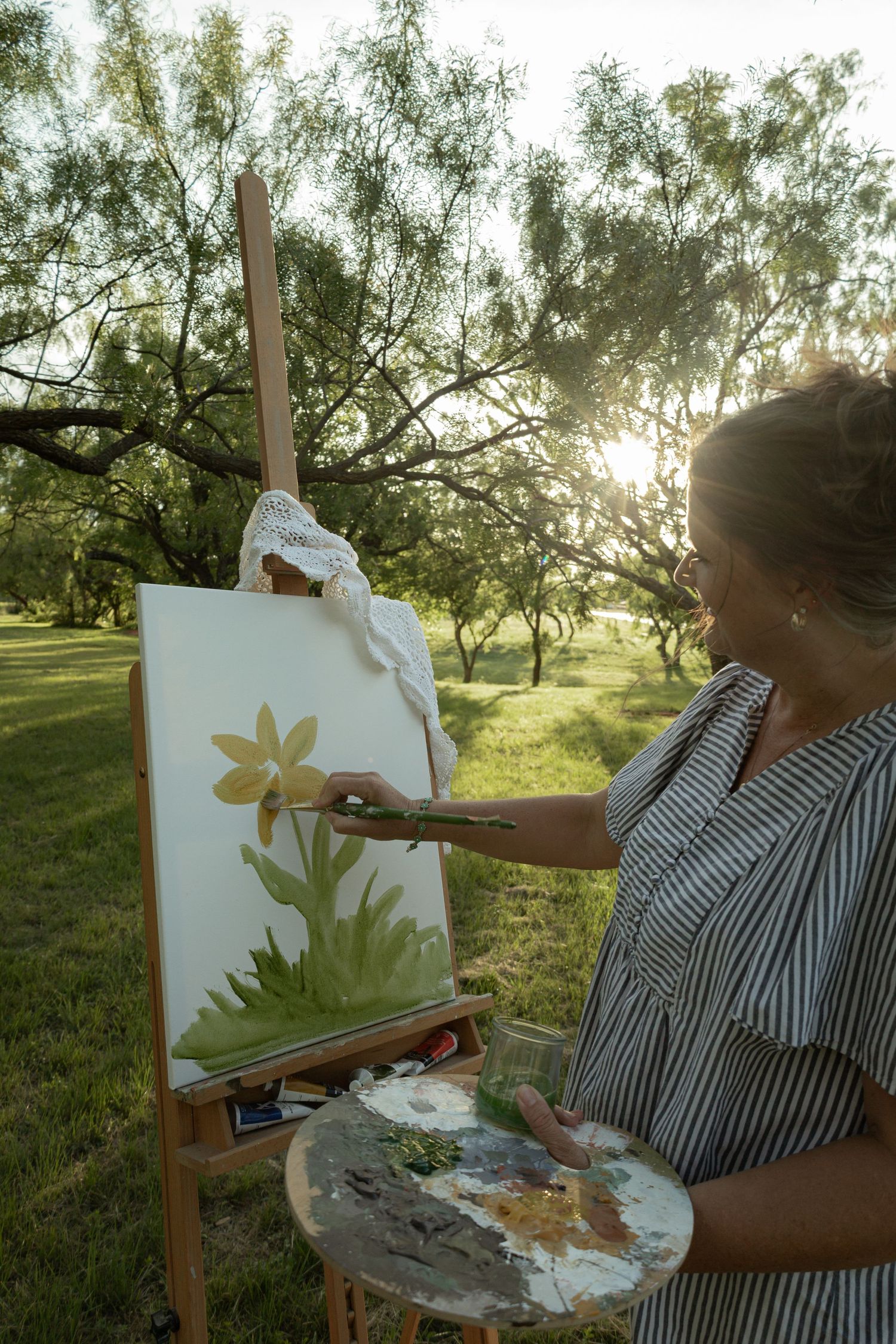 Woman painting flowers on canvas at an outdoor easel in a sunlit park, holding a palette and brush.