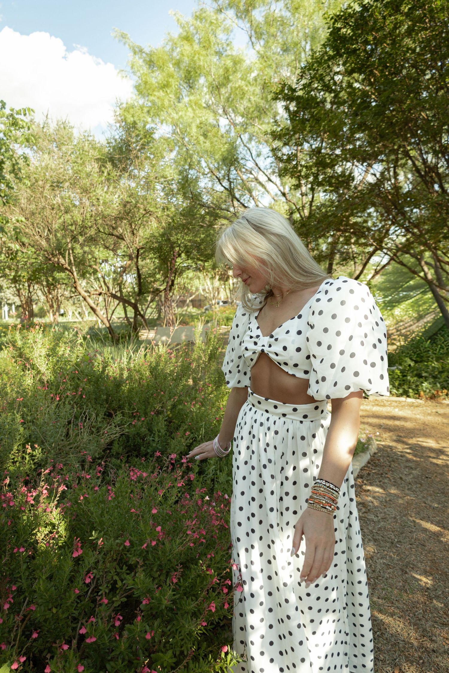 Woman in white polka dot midi dress touching pink flowers in a lush green park on a sunny day.