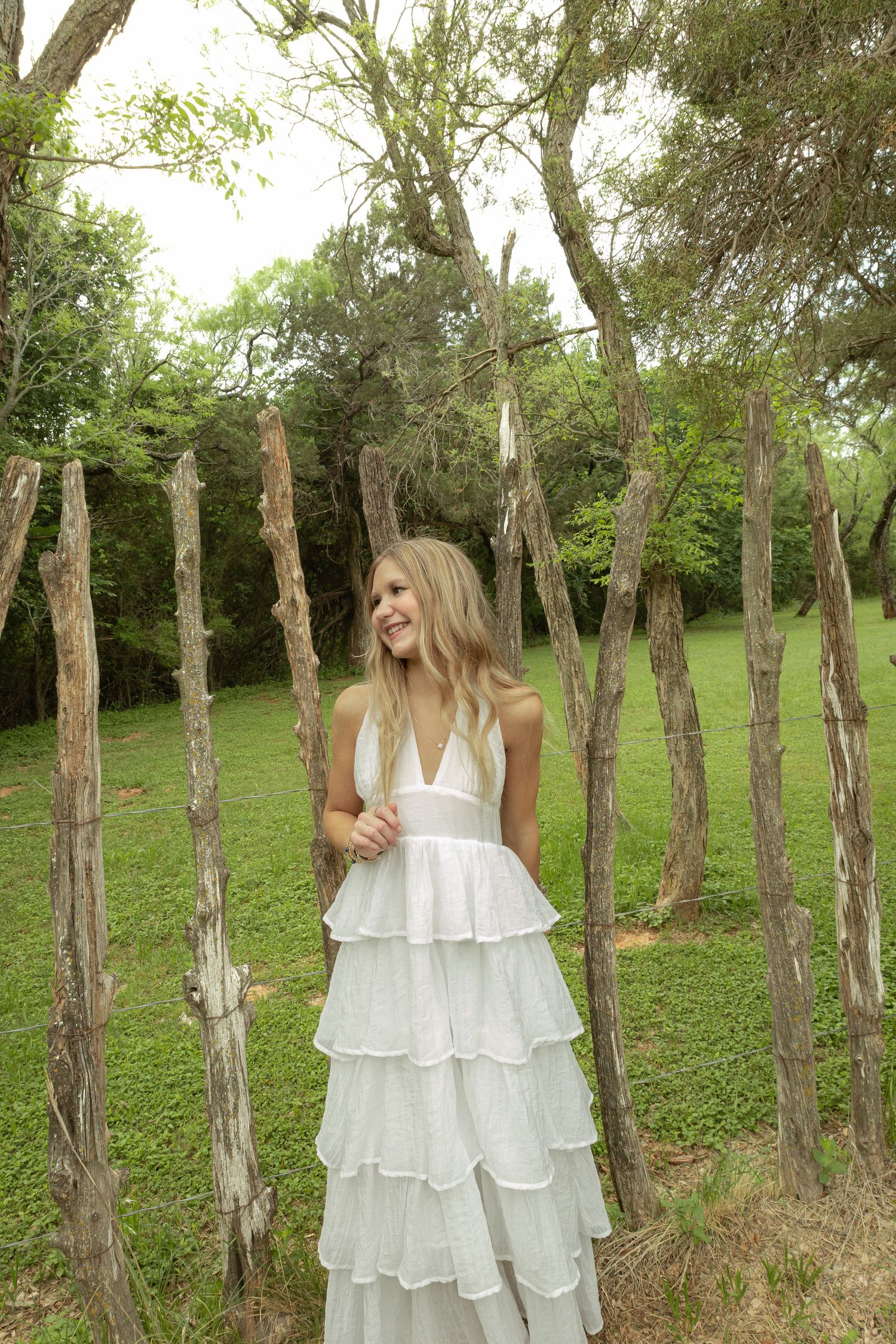 A smiling blonde woman in a white tiered ruffle maxi dress standing among rustic wooden posts in a lush green garden.