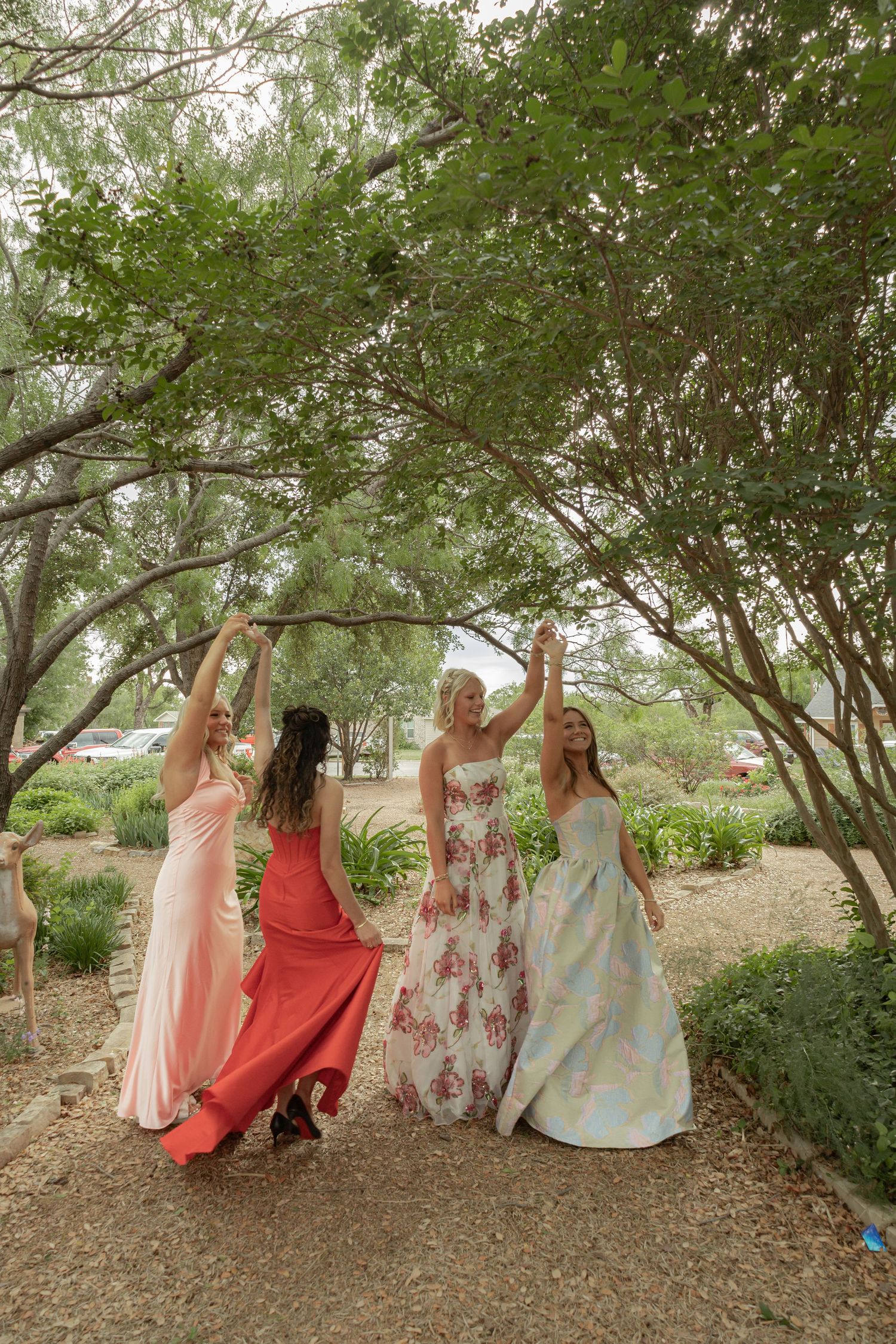 Four women in elegant formal gowns laughing and reaching up to touch tree branches in a garden setting.