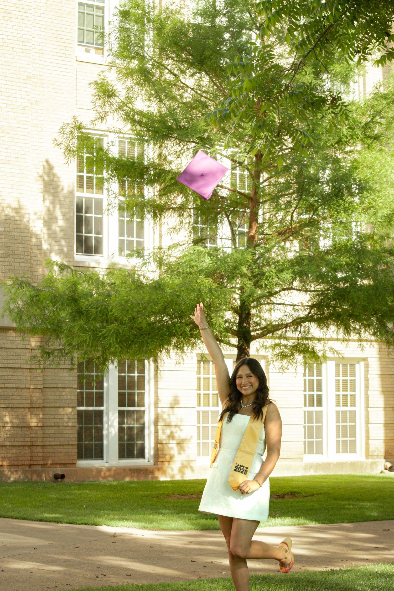 Young woman in white dress tossing pink graduation cap in the air outdoors near a brick university building.