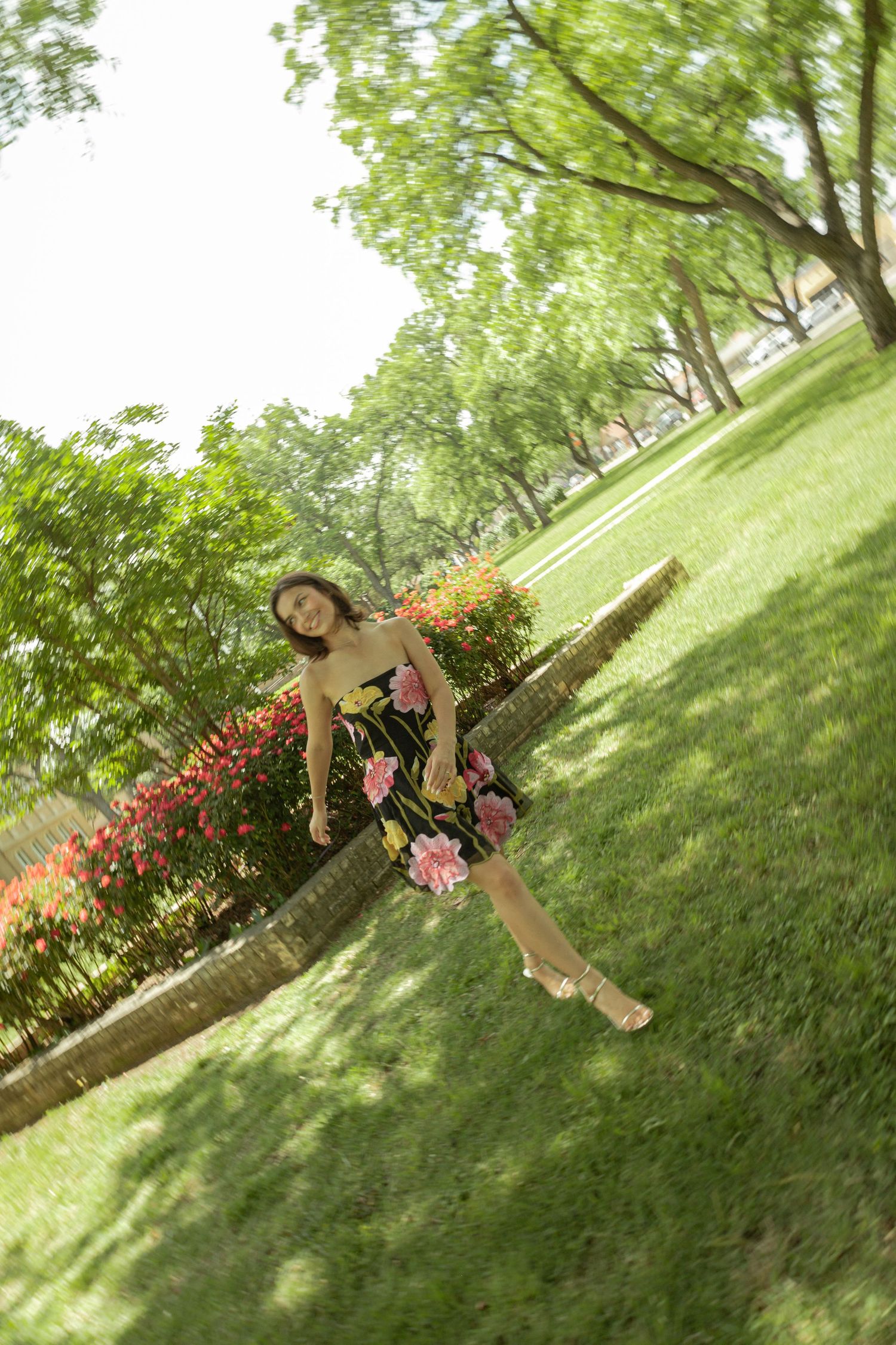 Young woman in floral dress posing playfully in a lush green park with trees and flowers.
