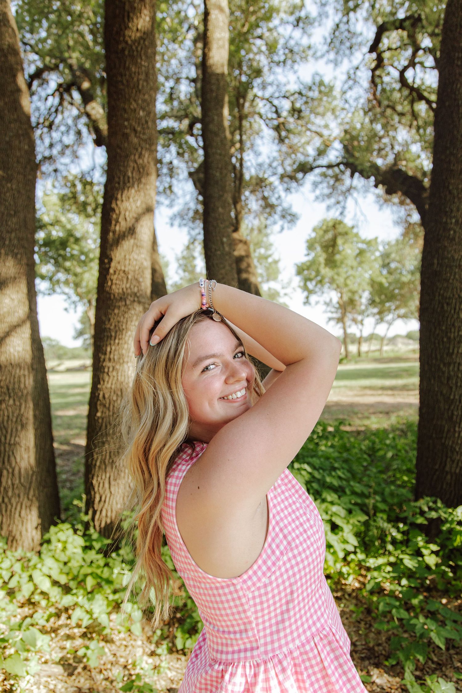 Young blonde woman in pink gingham dress smiling outdoors among tall trees in a lush green park setting.