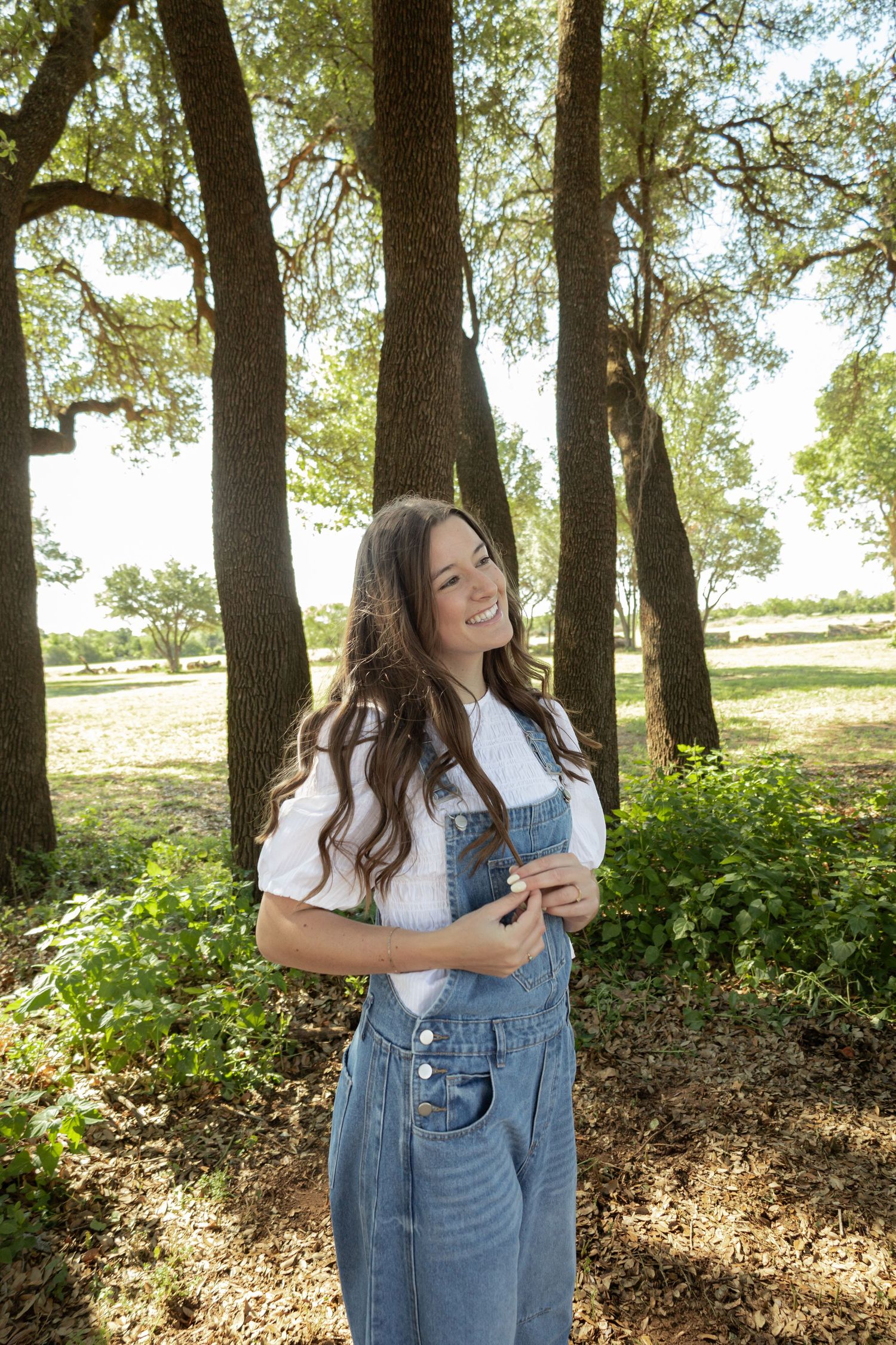 Smiling young woman in denim overalls and white shirt standing among trees in a sunny park.