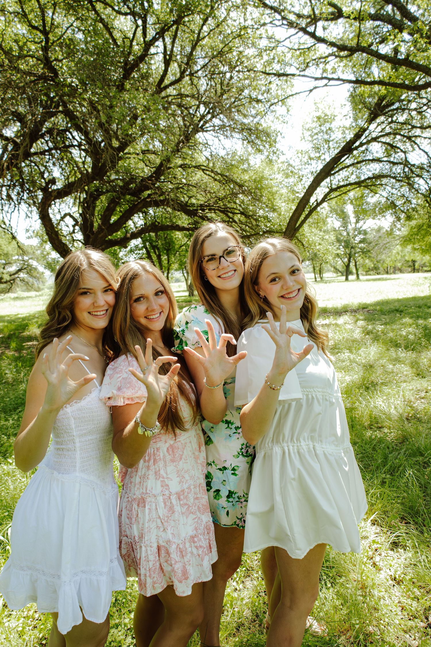 Four young women in summer dresses smile and pose together outdoors under green trees on a sunny day.