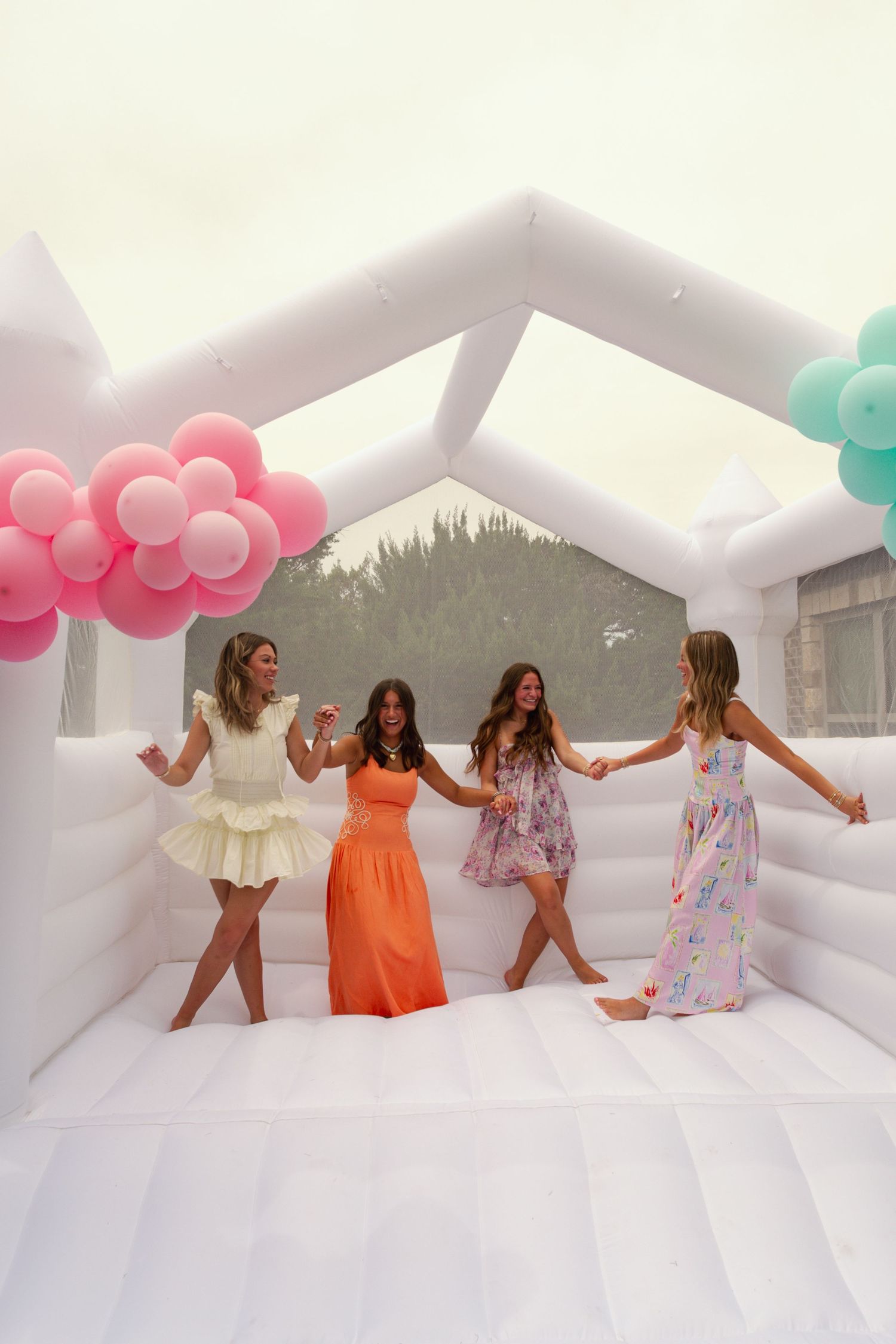 Four women in colorful dresses dancing in a white bounce house decorated with pink and mint green balloon clusters.