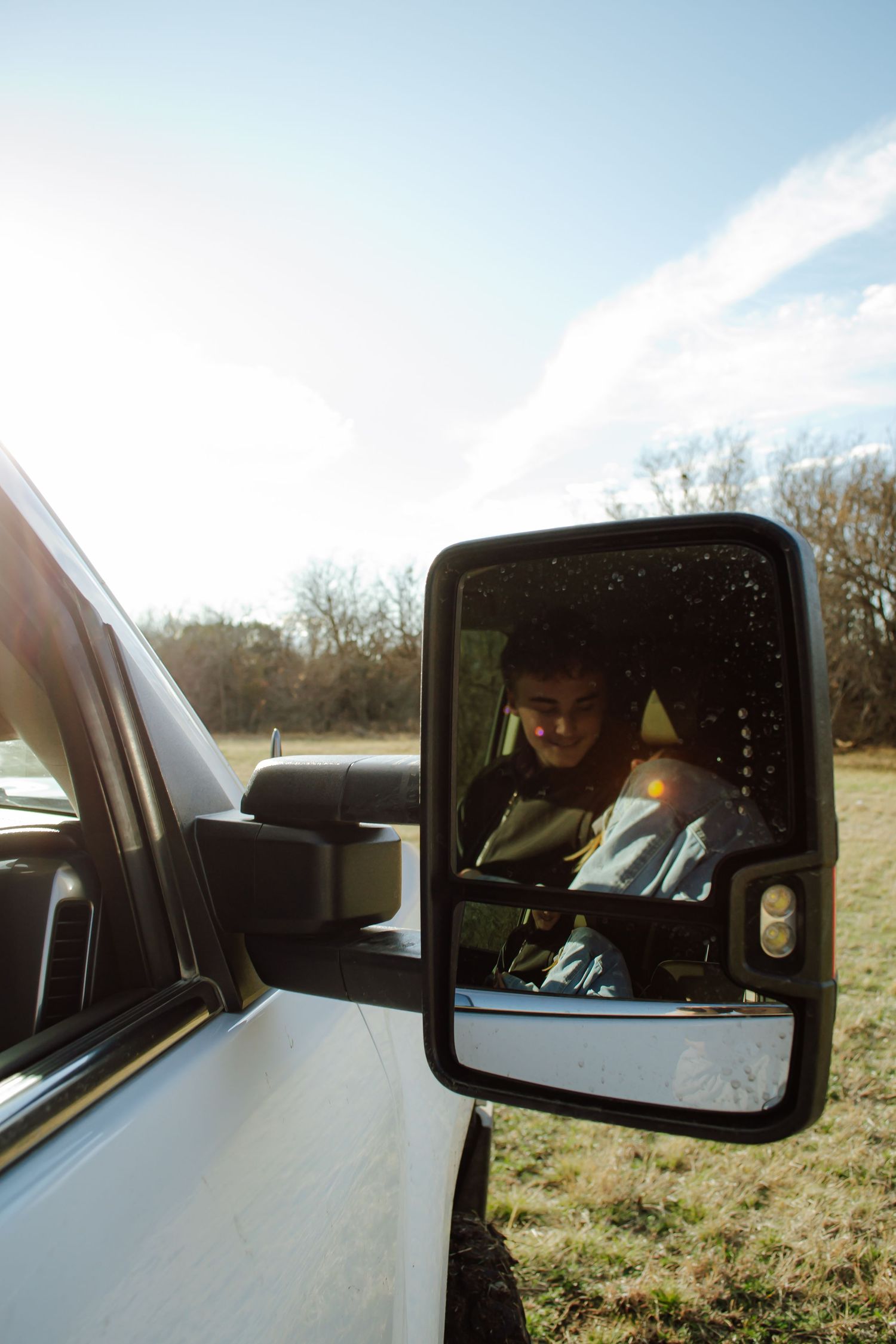 Man reflected in large white truck side mirror outdoors on a sunny day with trees and blue sky in background.