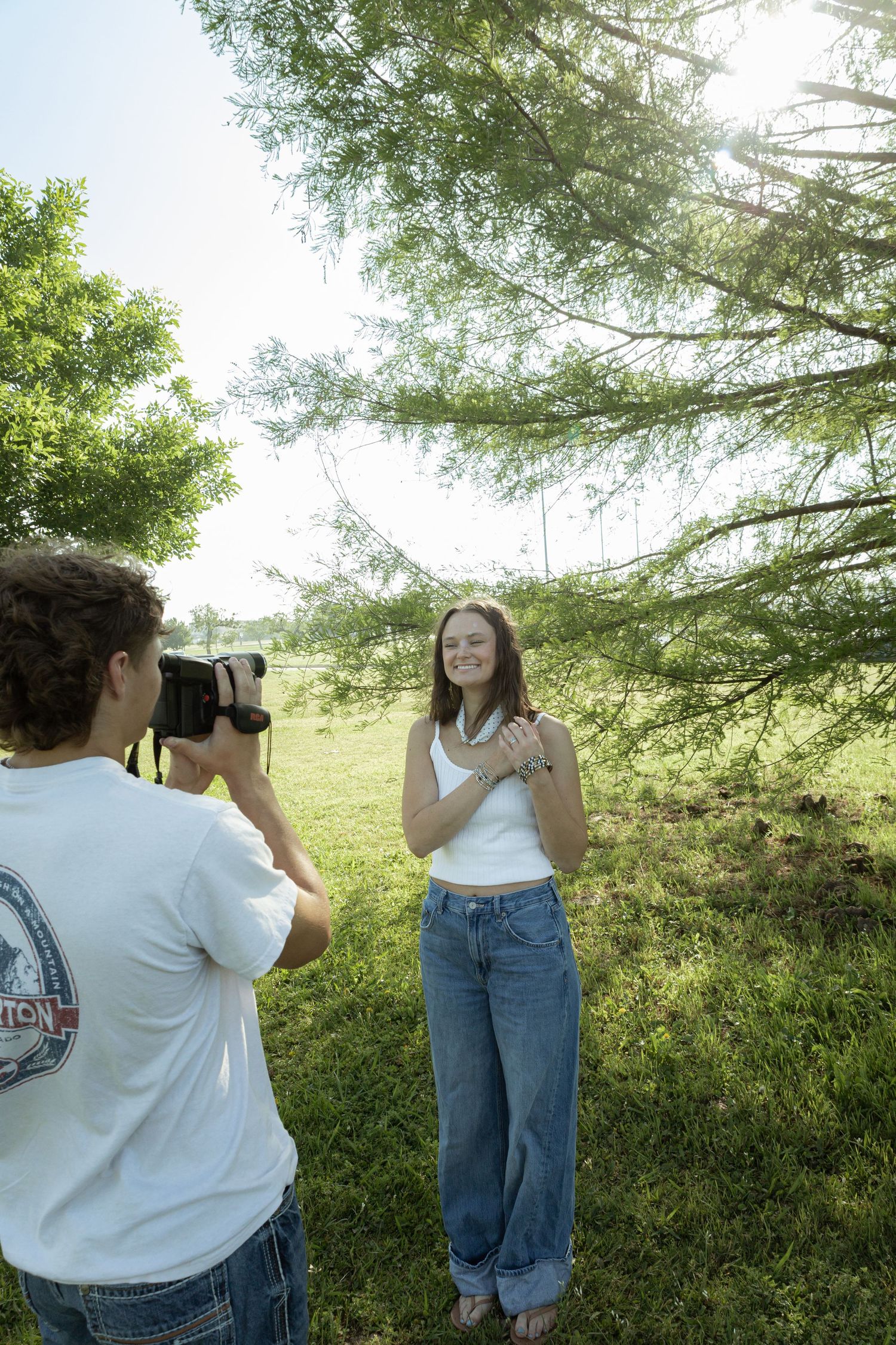 Young man photographing a smiling woman in jeans and white top outdoors near a pine tree on a sunny day.