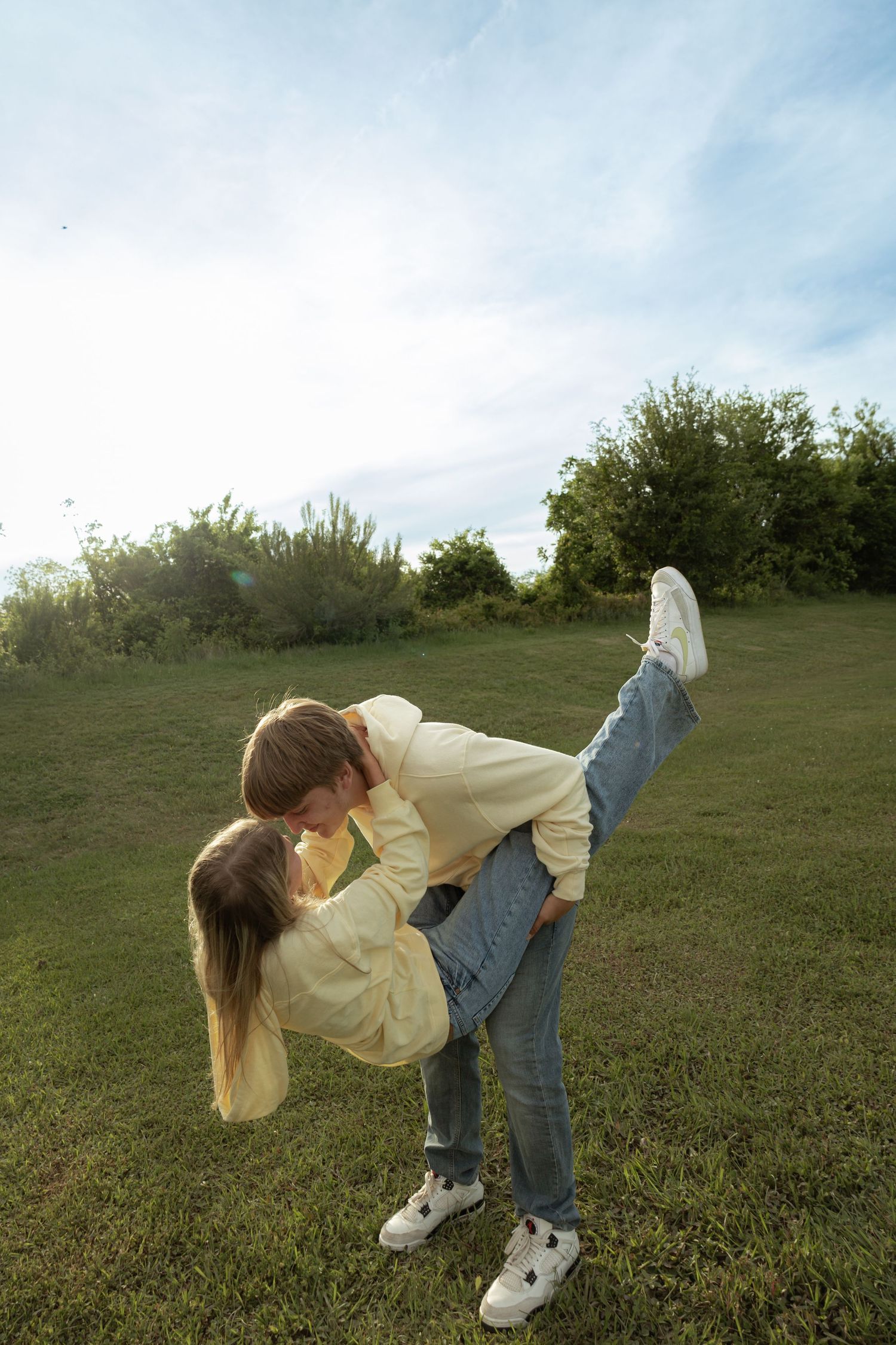 Couple sharing a romantic kiss outdoors on green grass, with trees and blue sky in the background on a sunny day.