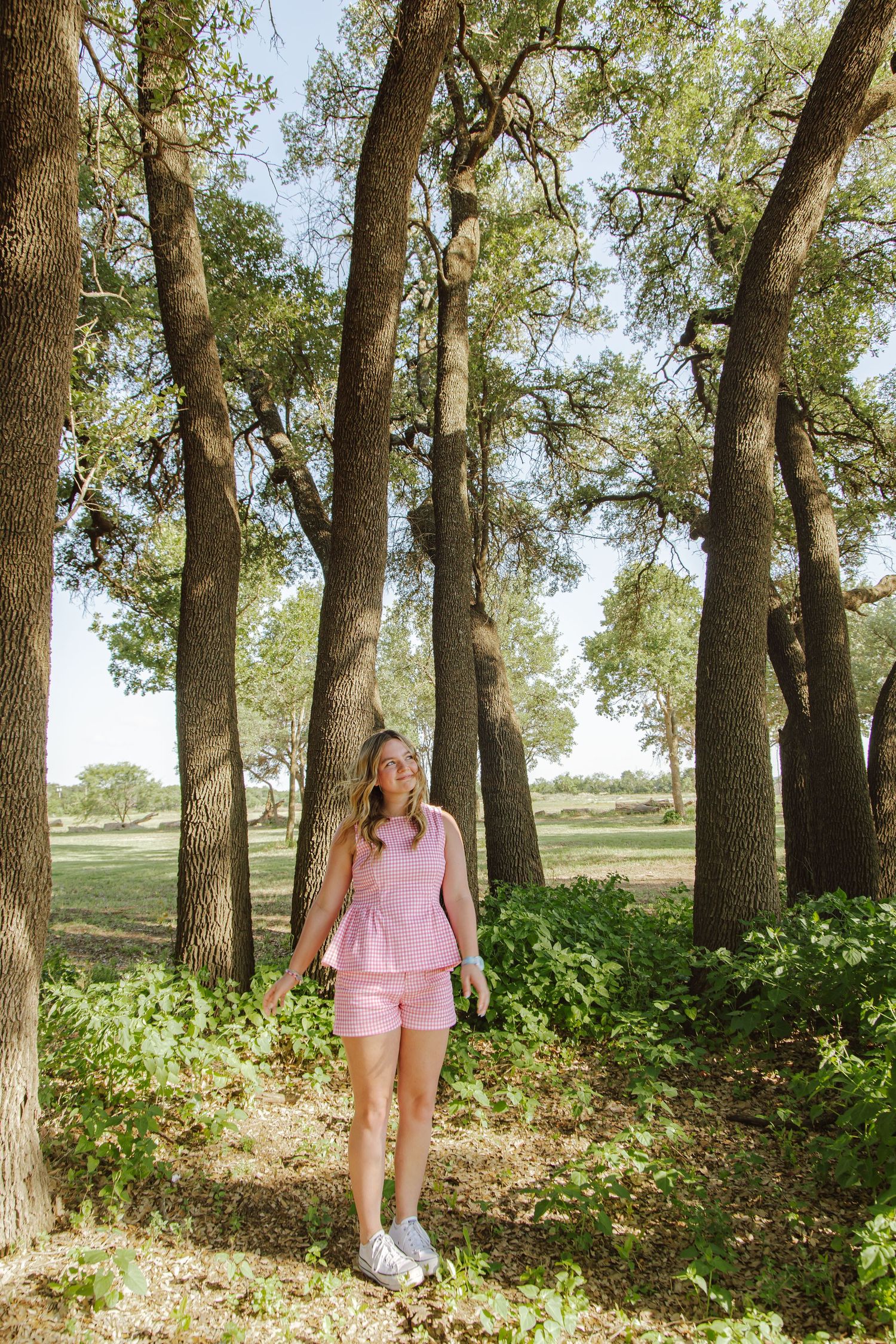 Woman in pink matching set standing among tall trees in a sunlit park, looking up at the canopy above.