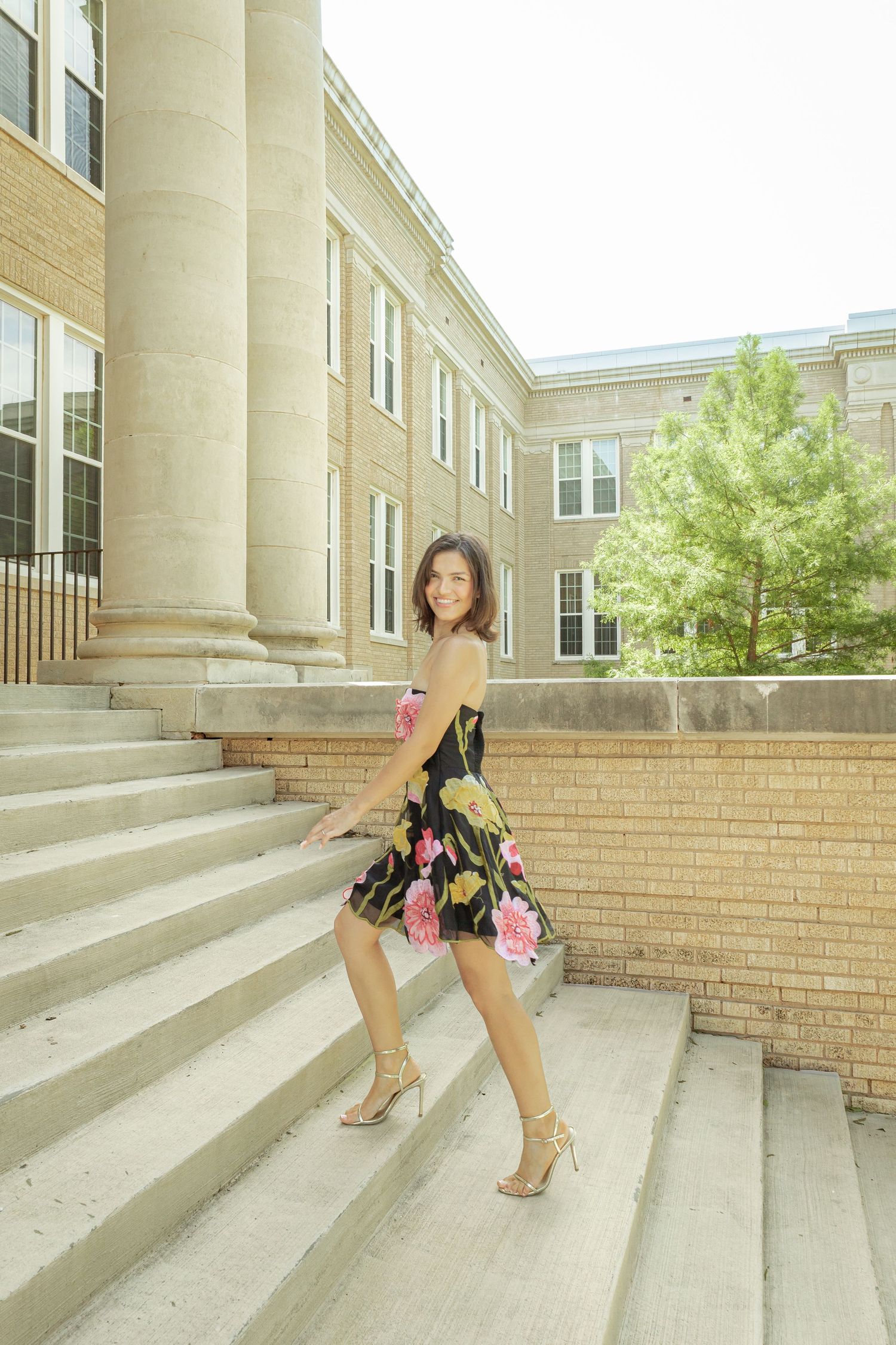 Young woman in floral dress and heels posing on stone steps outside a brick building with large columns.