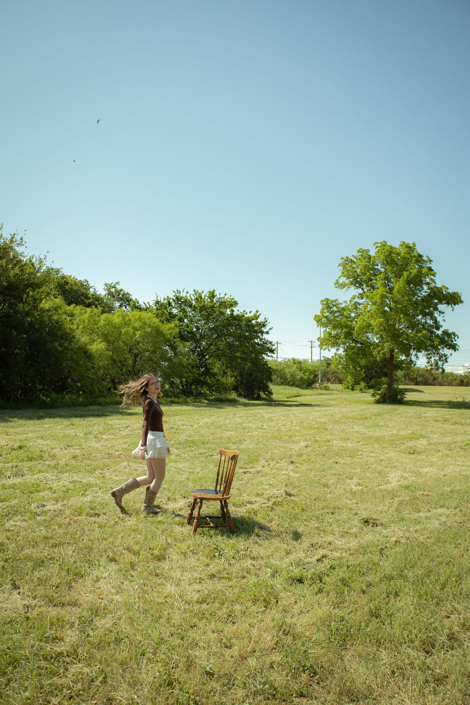 A young woman walks past a wooden chair in a sunny green meadow surrounded by trees under a clear blue sky.