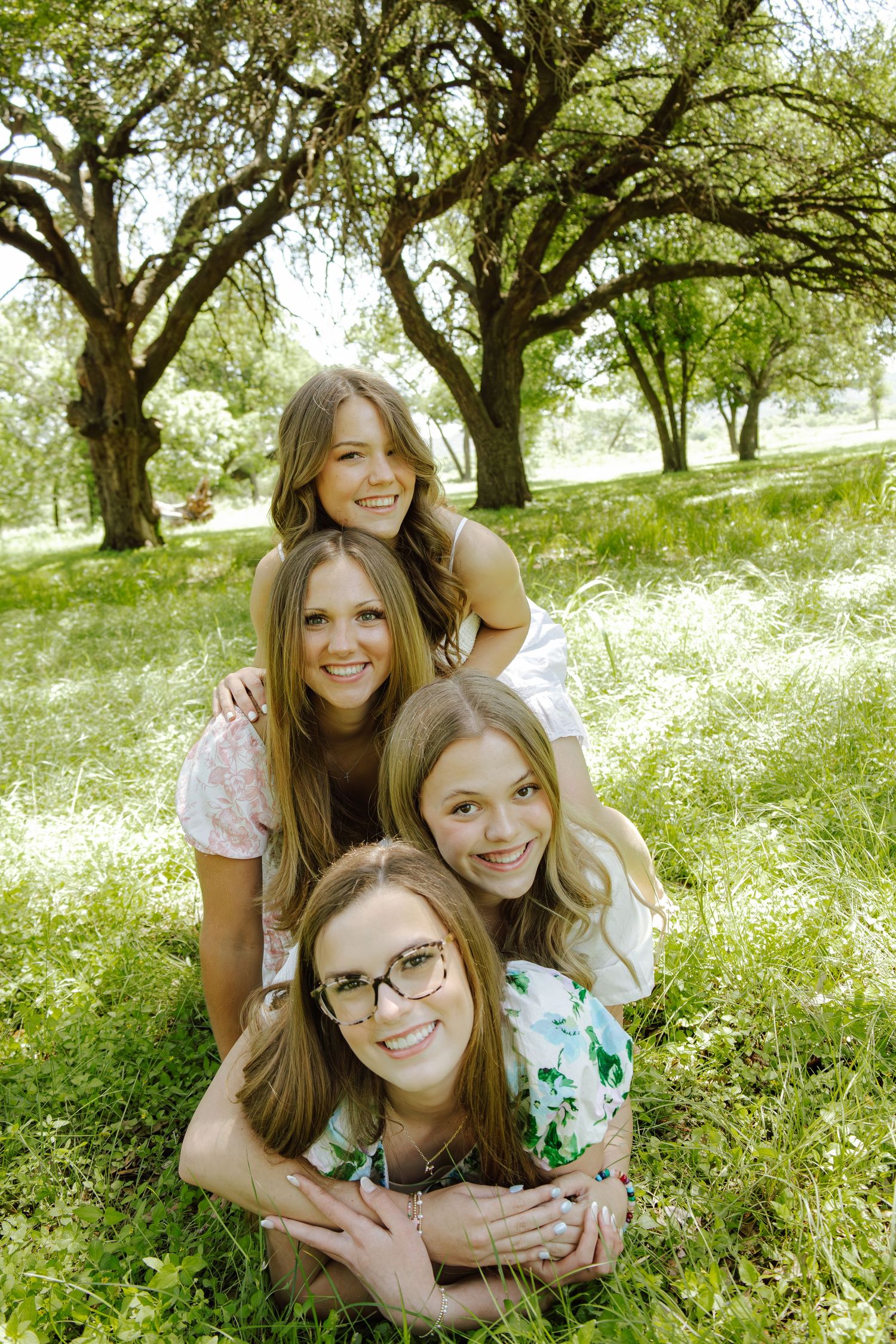 Four smiling young women stacked in a playful pyramid pose on green grass surrounded by trees outdoors.