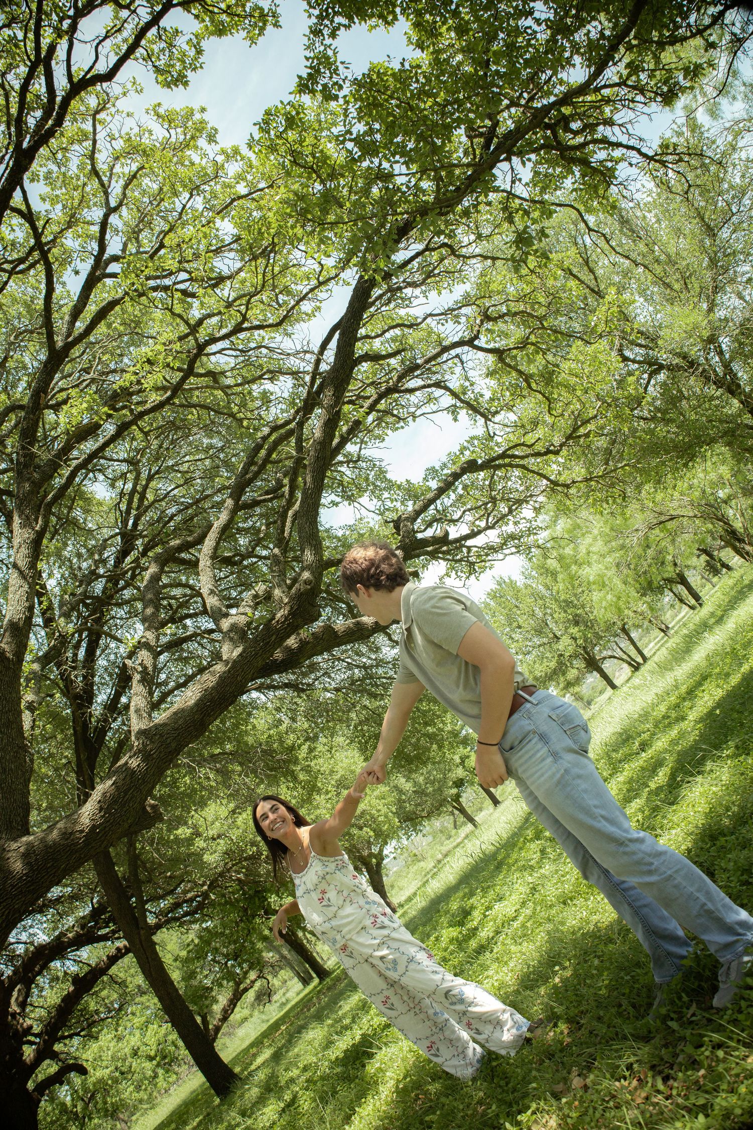 A man swings a young girl by her hands in a lush green park beneath sprawling oak trees on a sunny day.