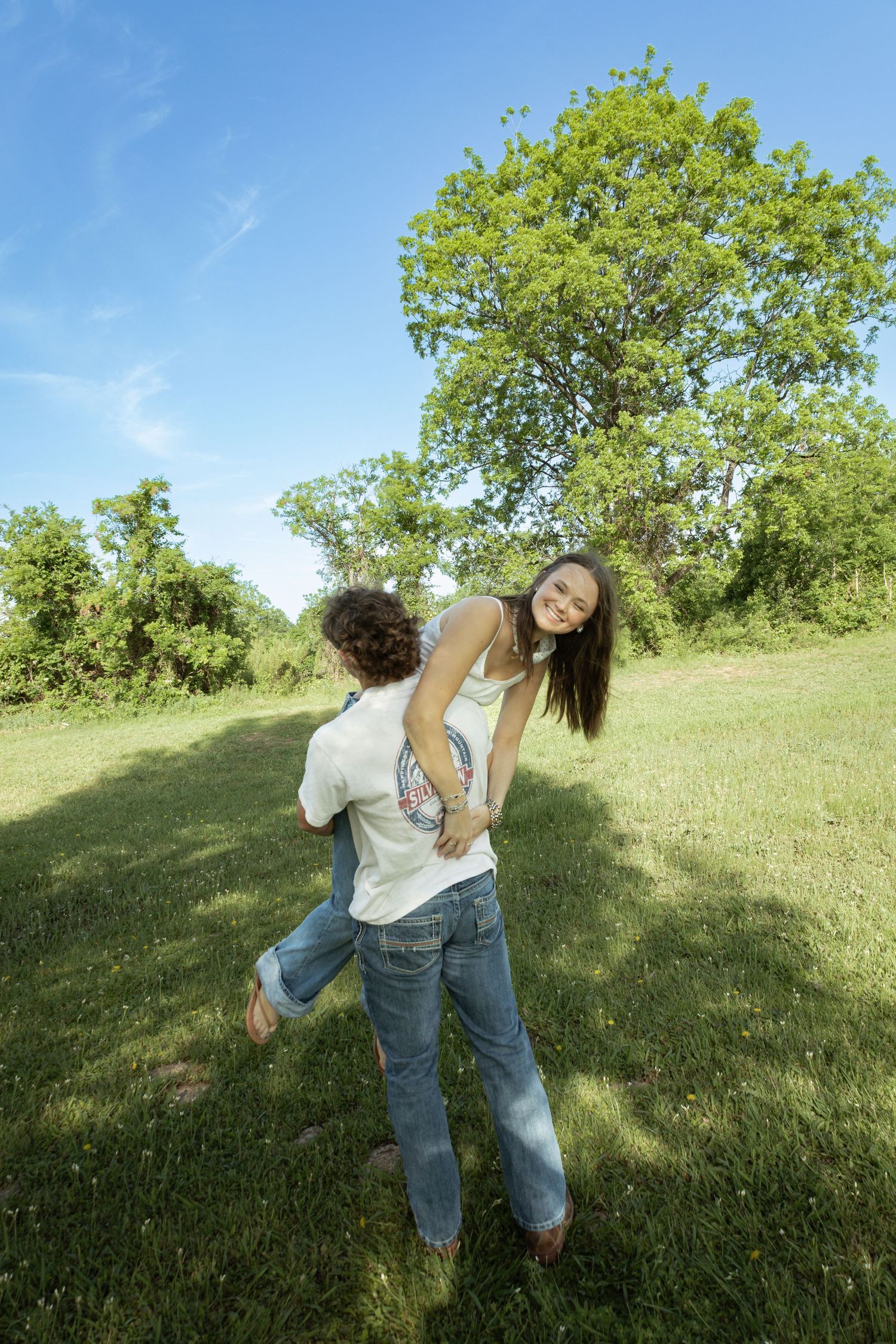 Two young women playfully dancing together on green grass in a sunny park with lush trees and blue sky.