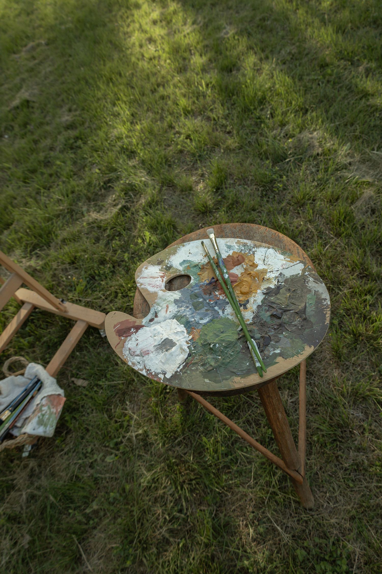 Artist's wooden paint palette with brushes on a tripod stand outdoors on green grass in natural light.