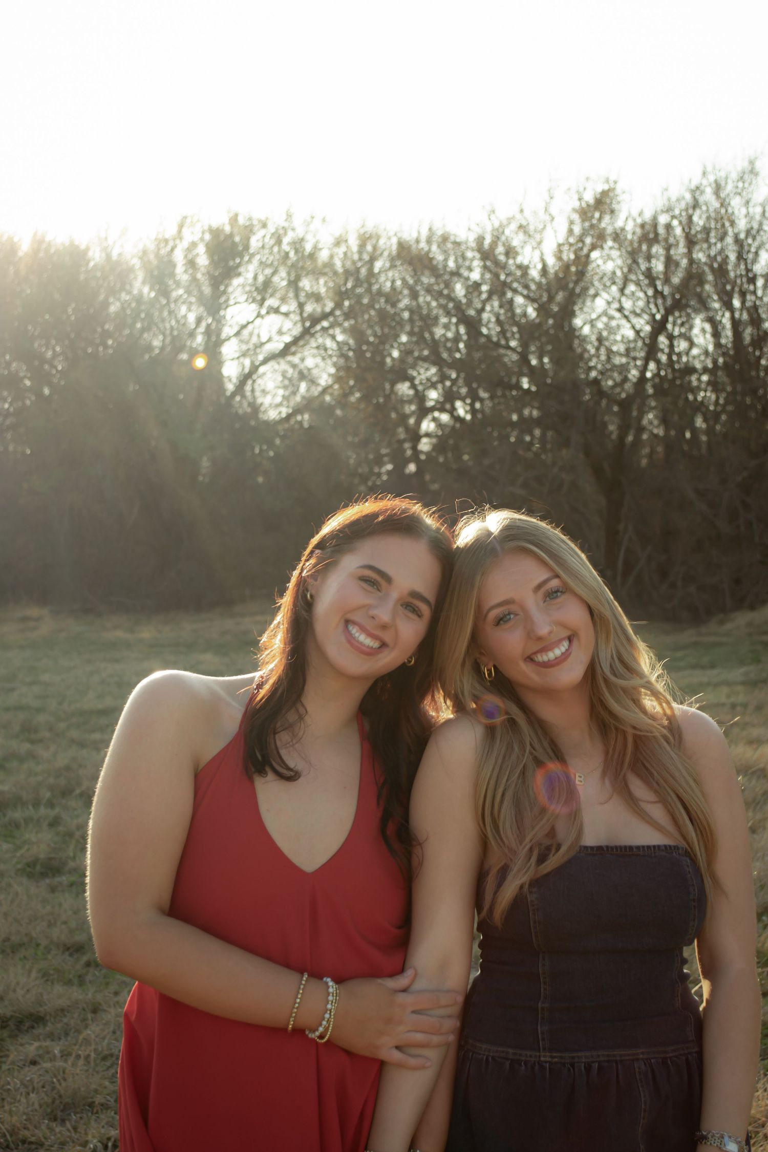 Two smiling young women in red and black dresses pose together in a sunlit outdoor field.