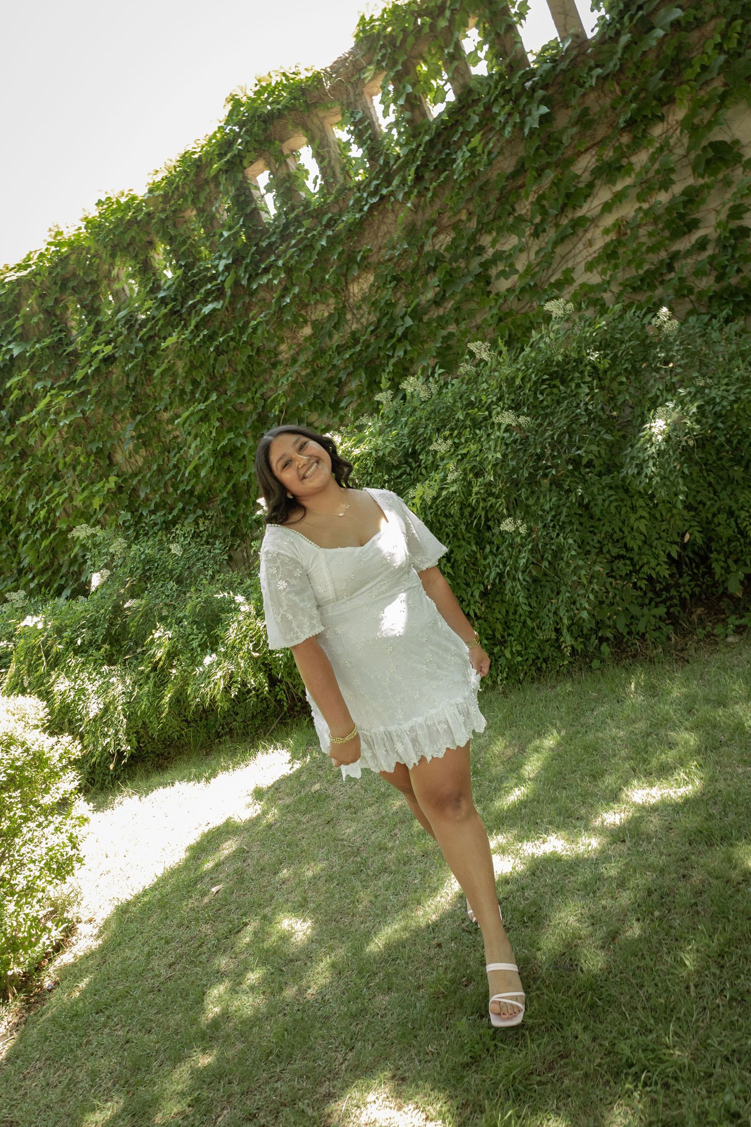 Young woman in white lace mini dress poses on grass beside ivy-covered brick wall on sunny day.
