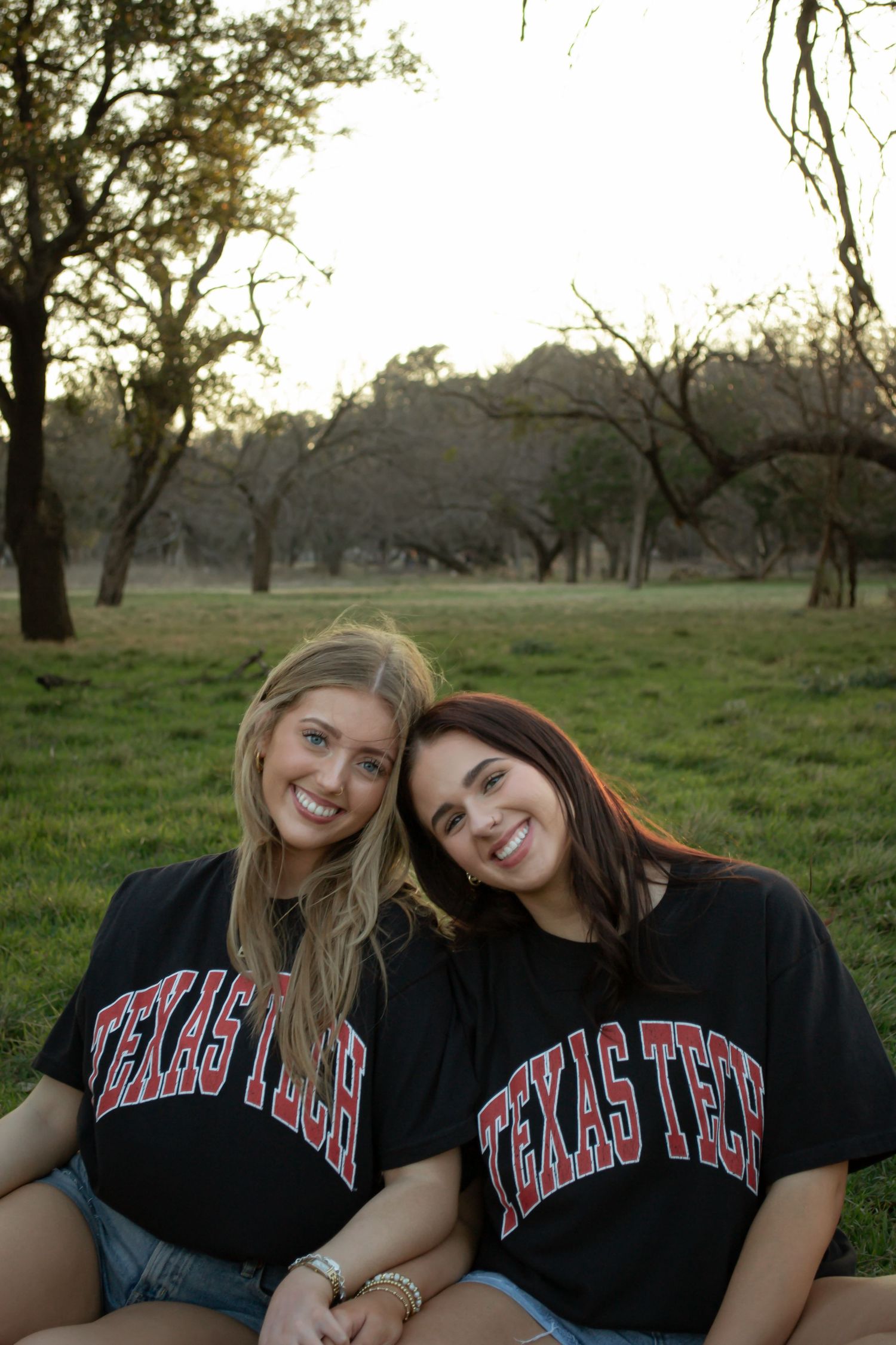 Two smiling women wearing matching black Texas Tech t-shirts sitting outdoors on green grass with trees in background.