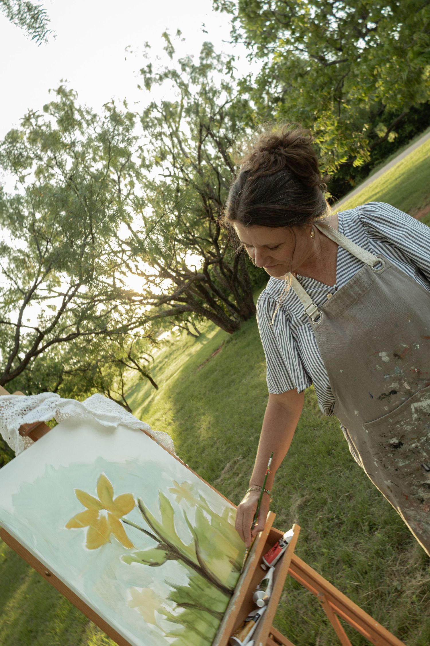 Woman painting yellow flowers on canvas at an outdoor easel in a sunny green park setting.