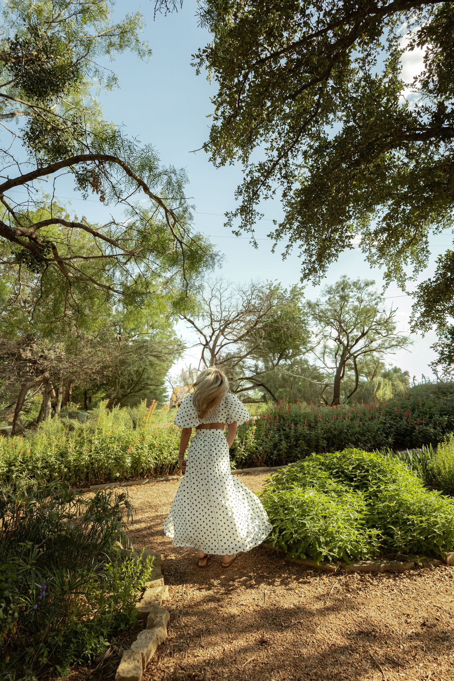 Woman in white polka dot dress and straw hat walking through a lush green garden path on a sunny day.