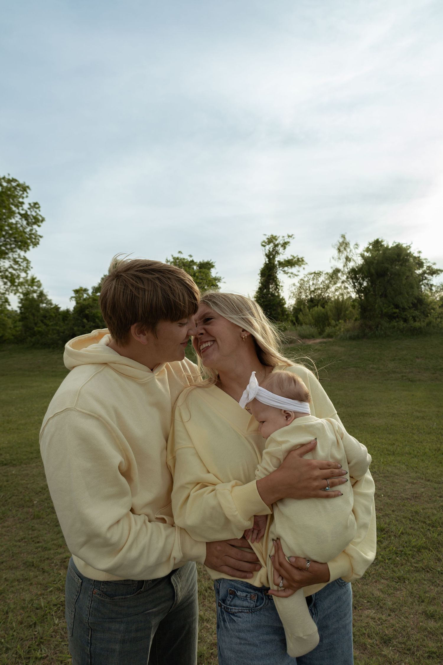Young couple in matching cream hoodies smiling at each other while holding their newborn baby outdoors.