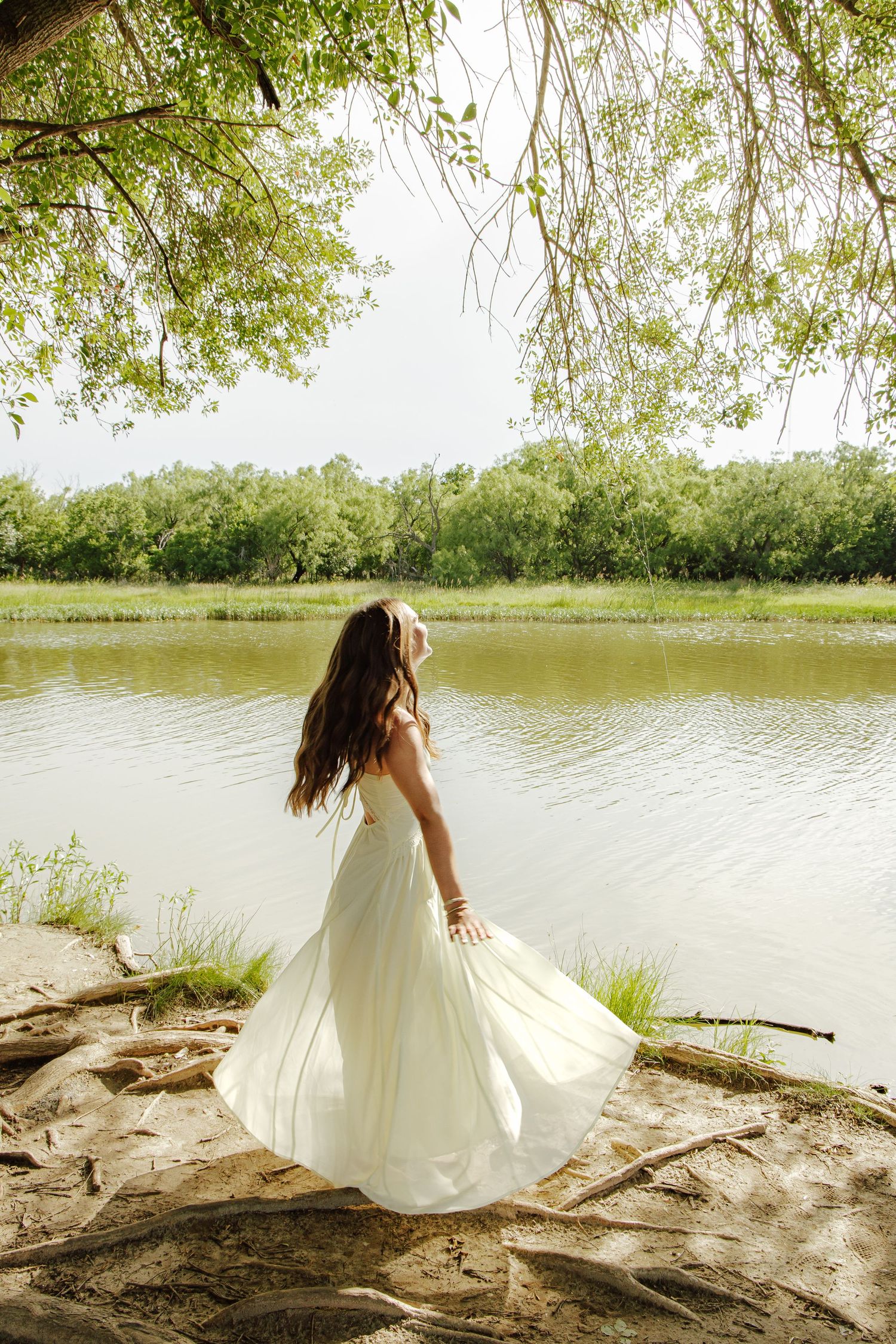 Young woman in flowing white dress twirling by a calm river with lush green trees in background.
