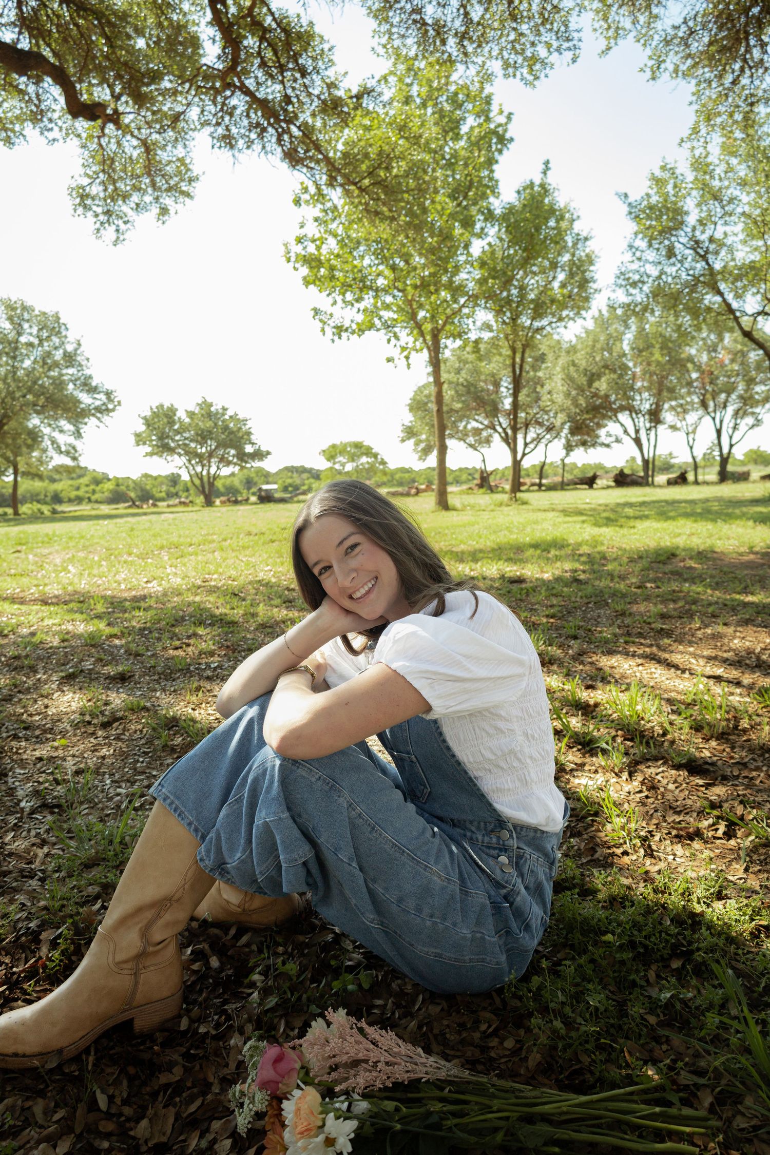 Young woman in denim overalls and boots sitting on grass in a sunny park, smiling cheerfully.