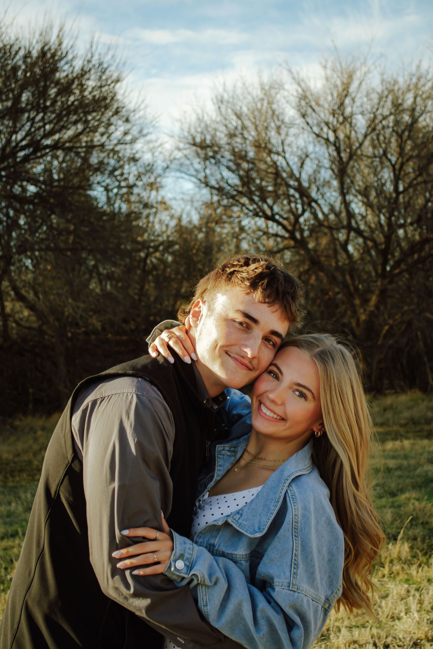 A young couple embracing outdoors, smiling warmly with bare trees and blue sky in the background.