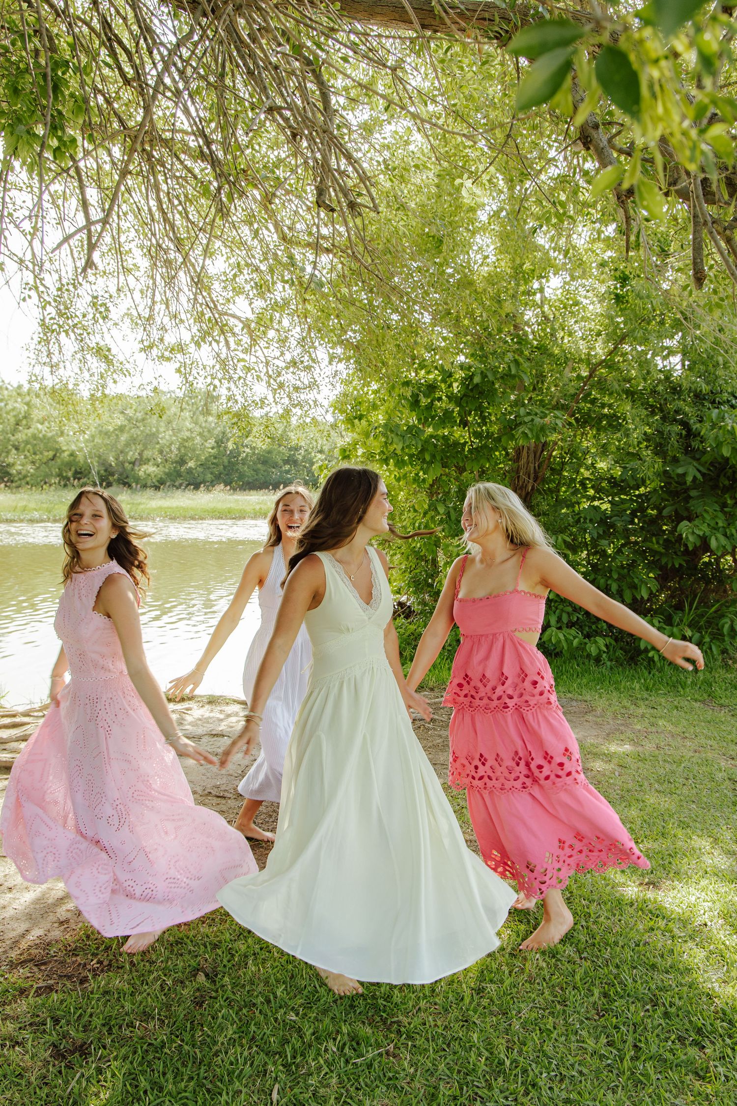Three women in pink and white dresses dancing joyfully outdoors near a lake surrounded by lush greenery.