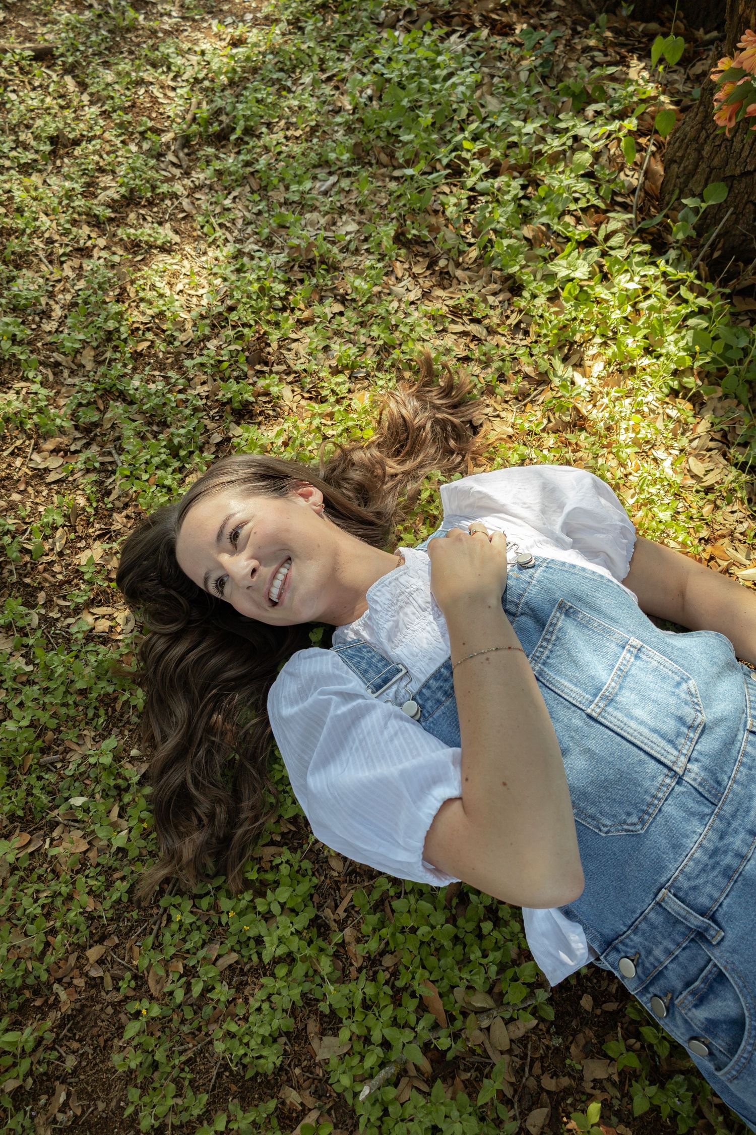 Young woman in denim overalls lying on grass, smiling joyfully in a sunlit park setting.