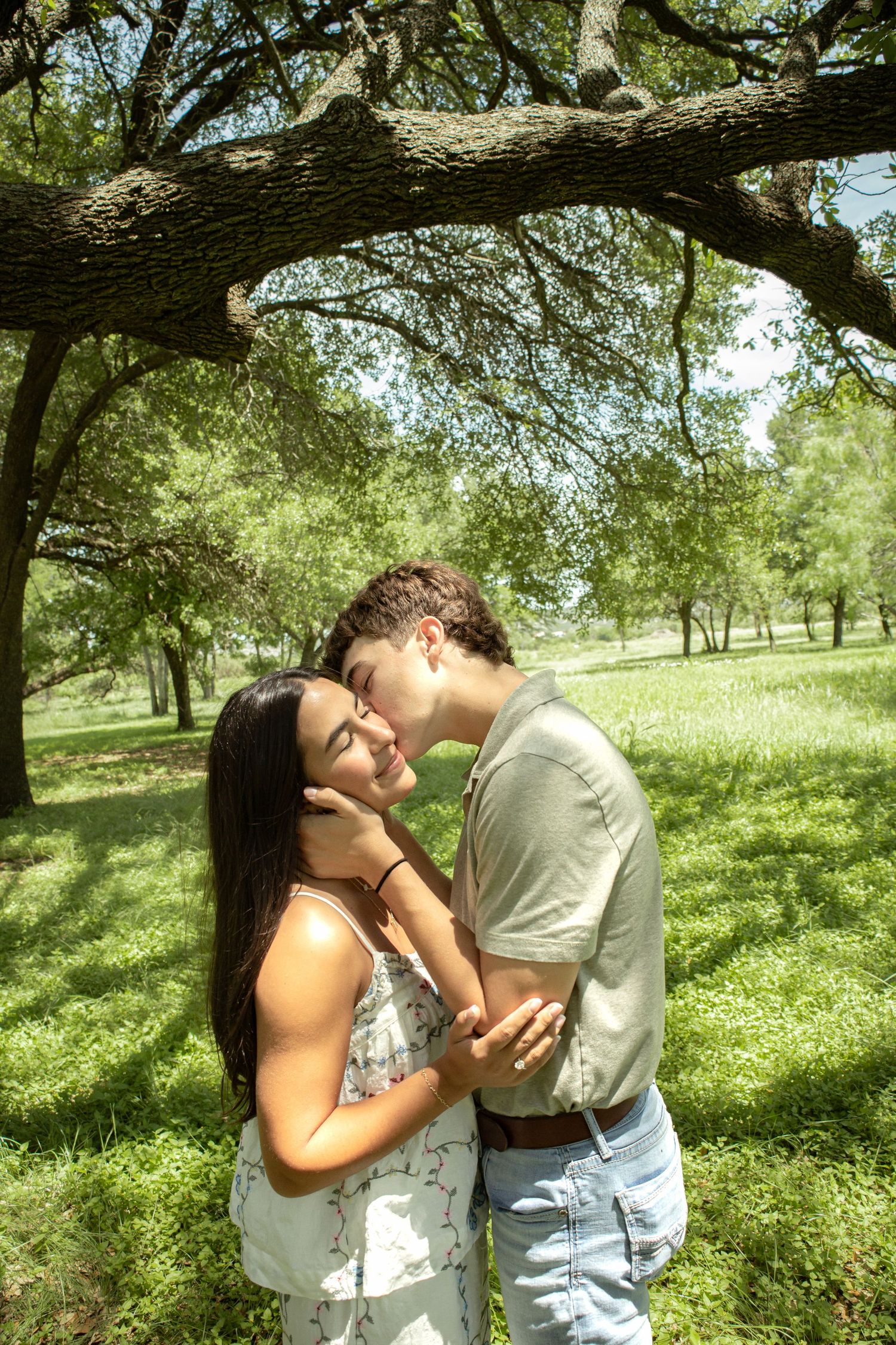 Young couple sharing a tender kiss in a lush green park under sprawling oak trees on a sunny day.