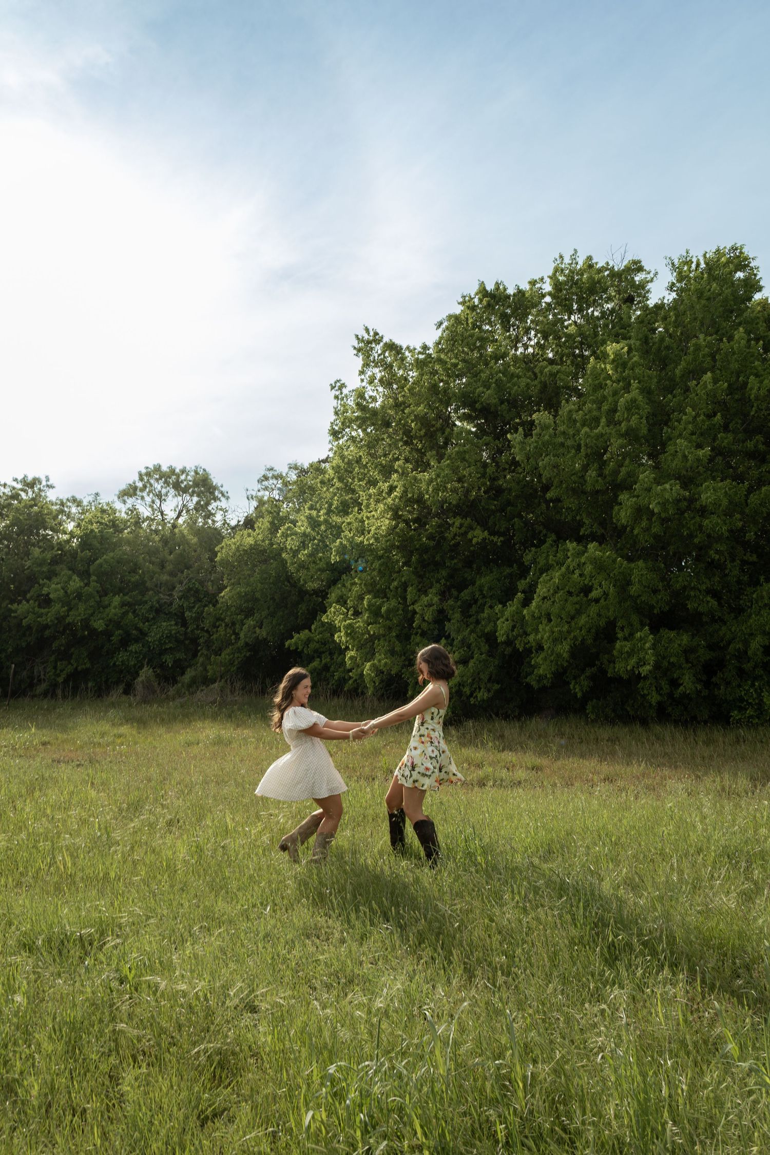 Two young women in summer dresses hold hands and spin in a lush green meadow with trees in the background.